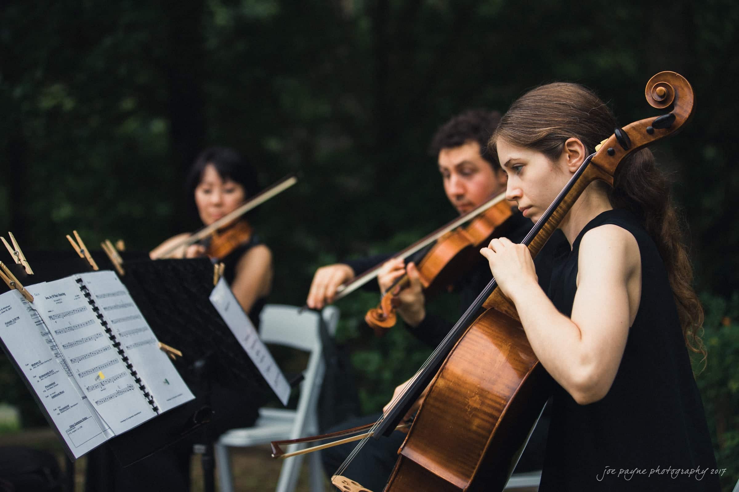 A string trio performs outdoors, featuring a cellist in the foreground and two violinists in the background. They are focused on their music, with sheet music visible on stands. Trees provide a natural backdrop.