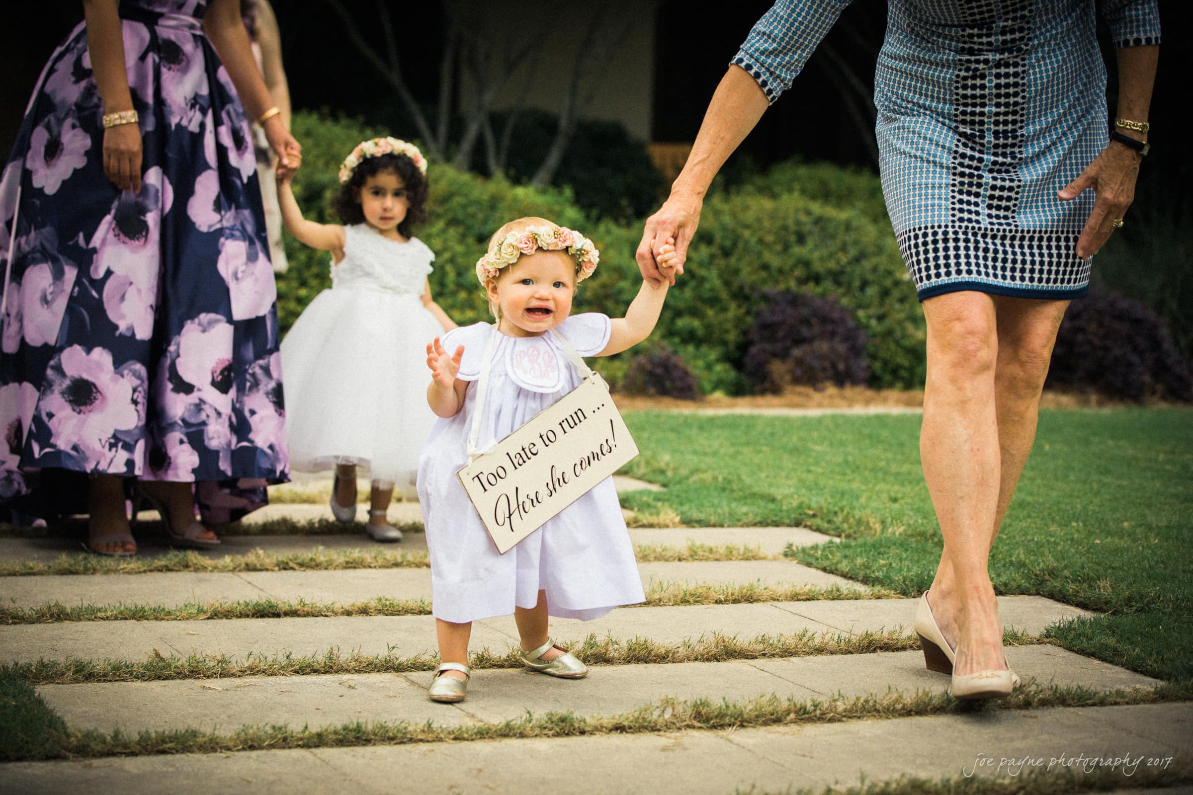 A baby wearing a flower crown and holding a sign that reads Too late to run... Here she comes! is walking with an adult. A young girl in a white dress follows behind. They are outdoors, with grass and shrubbery in the background.