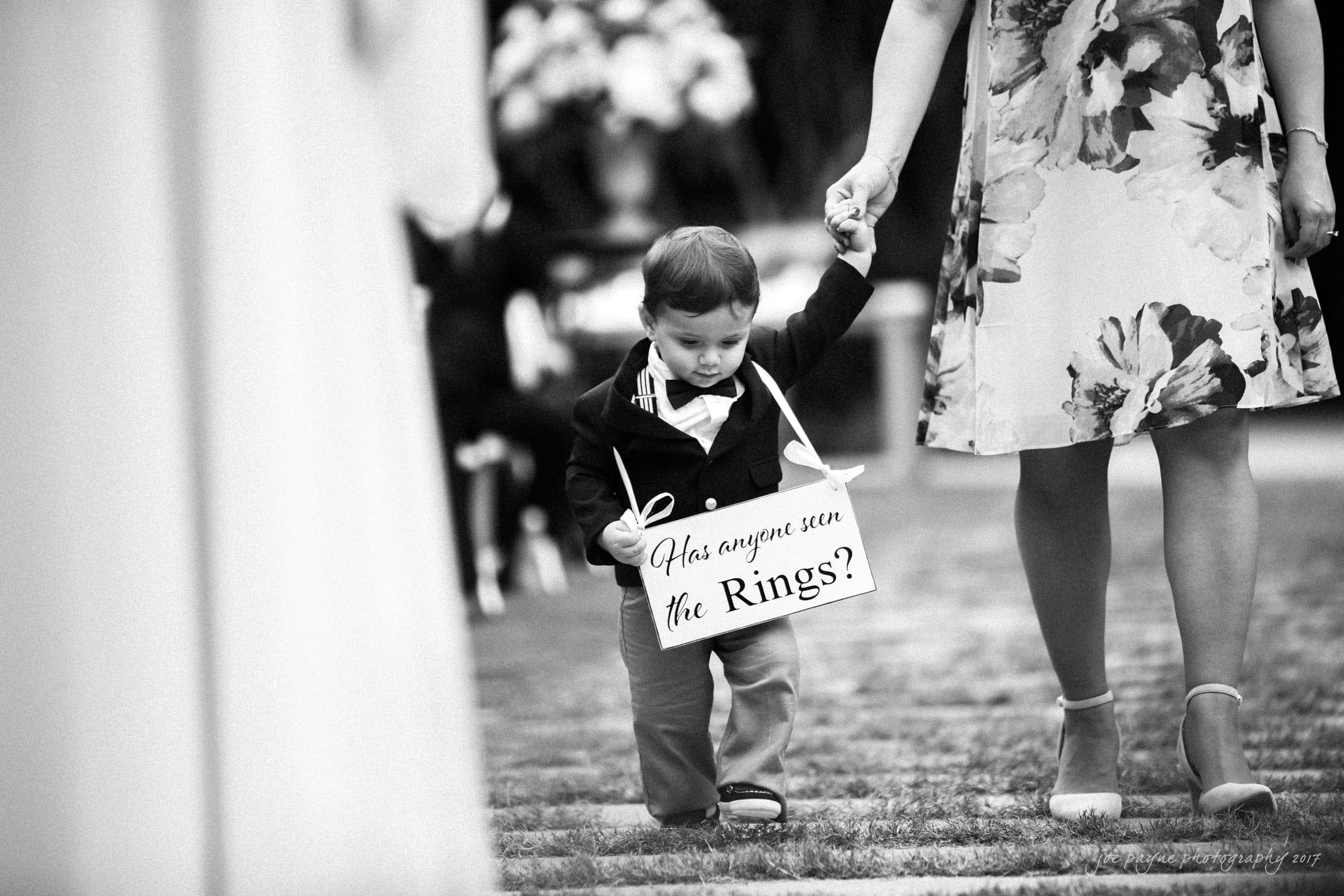A young child dressed in formal attire holds a womans hand while walking on grass. The child carries a sign that reads, Has anyone seen the rings? The scene appears to be at a wedding, with floral decorations in the background.