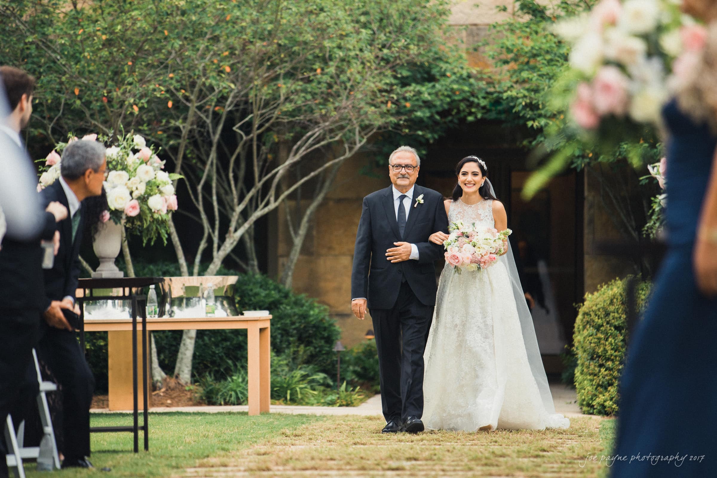 A bride in a white wedding gown walks down a grassy aisle with an older man in a black suit. They are surrounded by guests and floral arrangements under a clear sky. The setting is outdoors with trees and greenery.