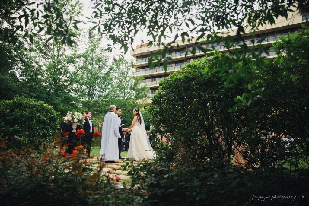 A bride and groom stand with an officiant during an outdoor wedding ceremony surrounded by greenery and flowers. Guests are gathered in the background near a multi-story building.