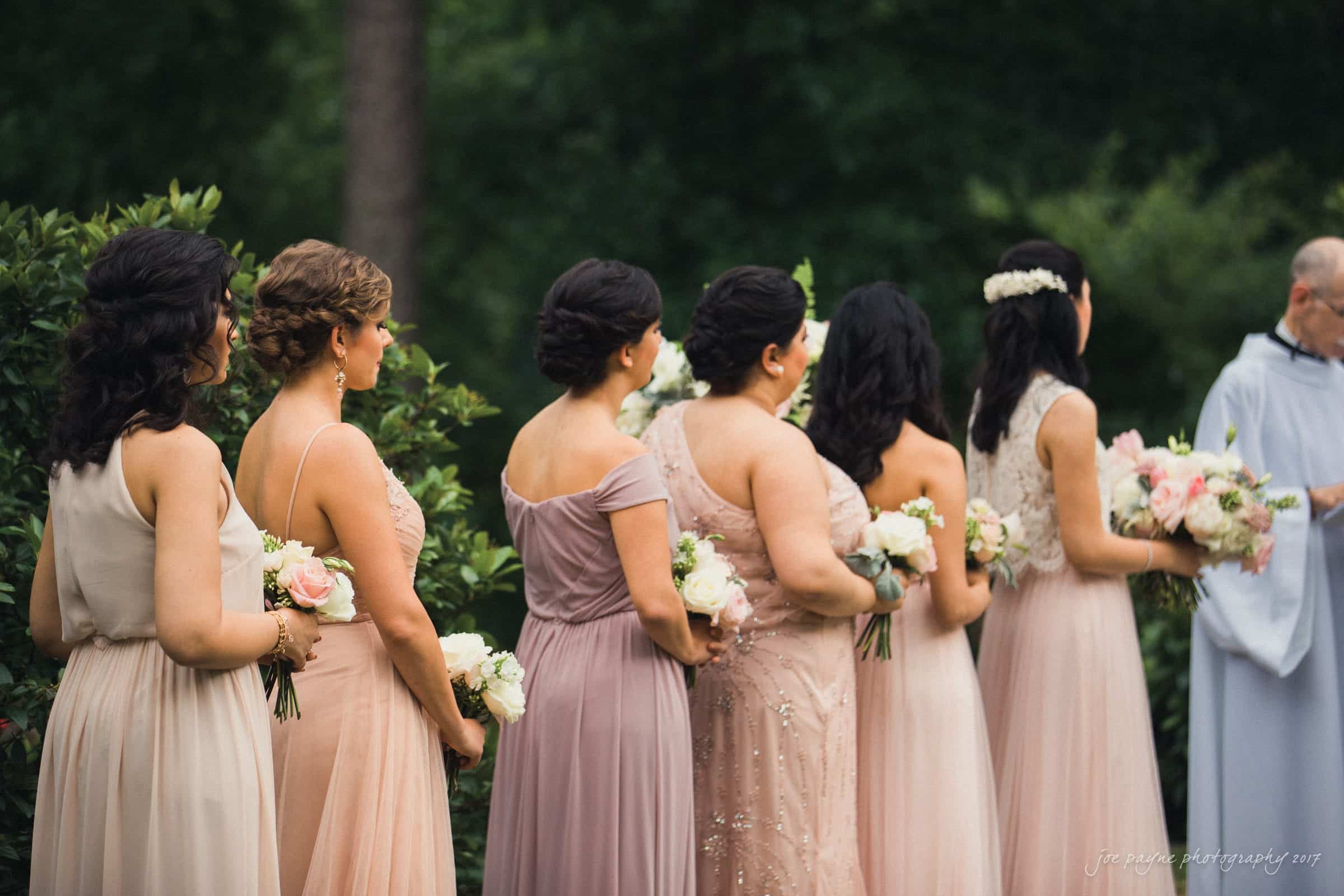 A group of bridesmaids in elegant, light pink dresses stand in a row outdoors, holding bouquets of flowers. They face away towards a person in a light blue outfit, set against a lush green background.
