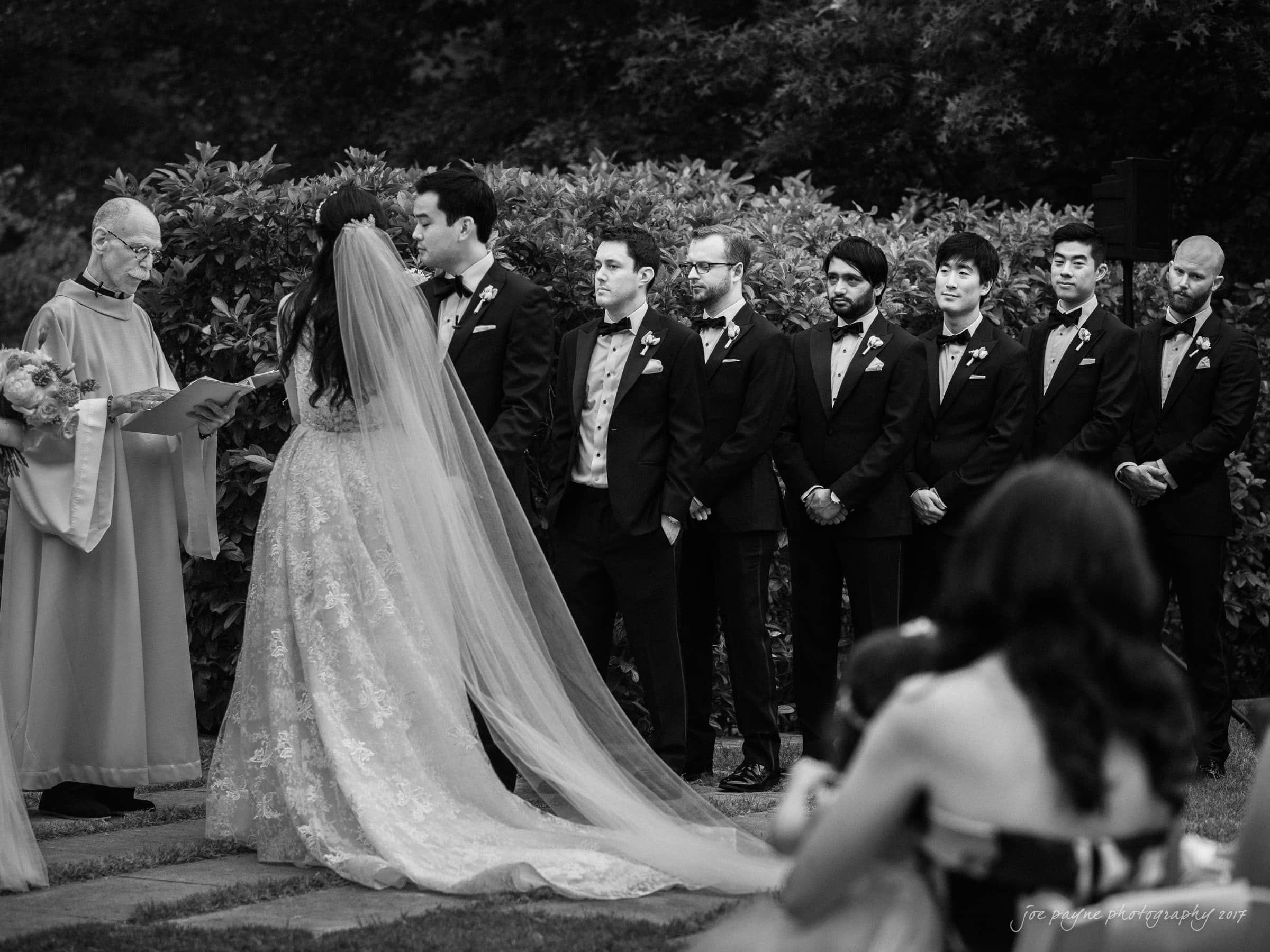 A black-and-white photo of a wedding ceremony. The bride and groom stand facing each other as the officiant reads. Several groomsmen stand lined up in the background, and guests are seated in the foreground.