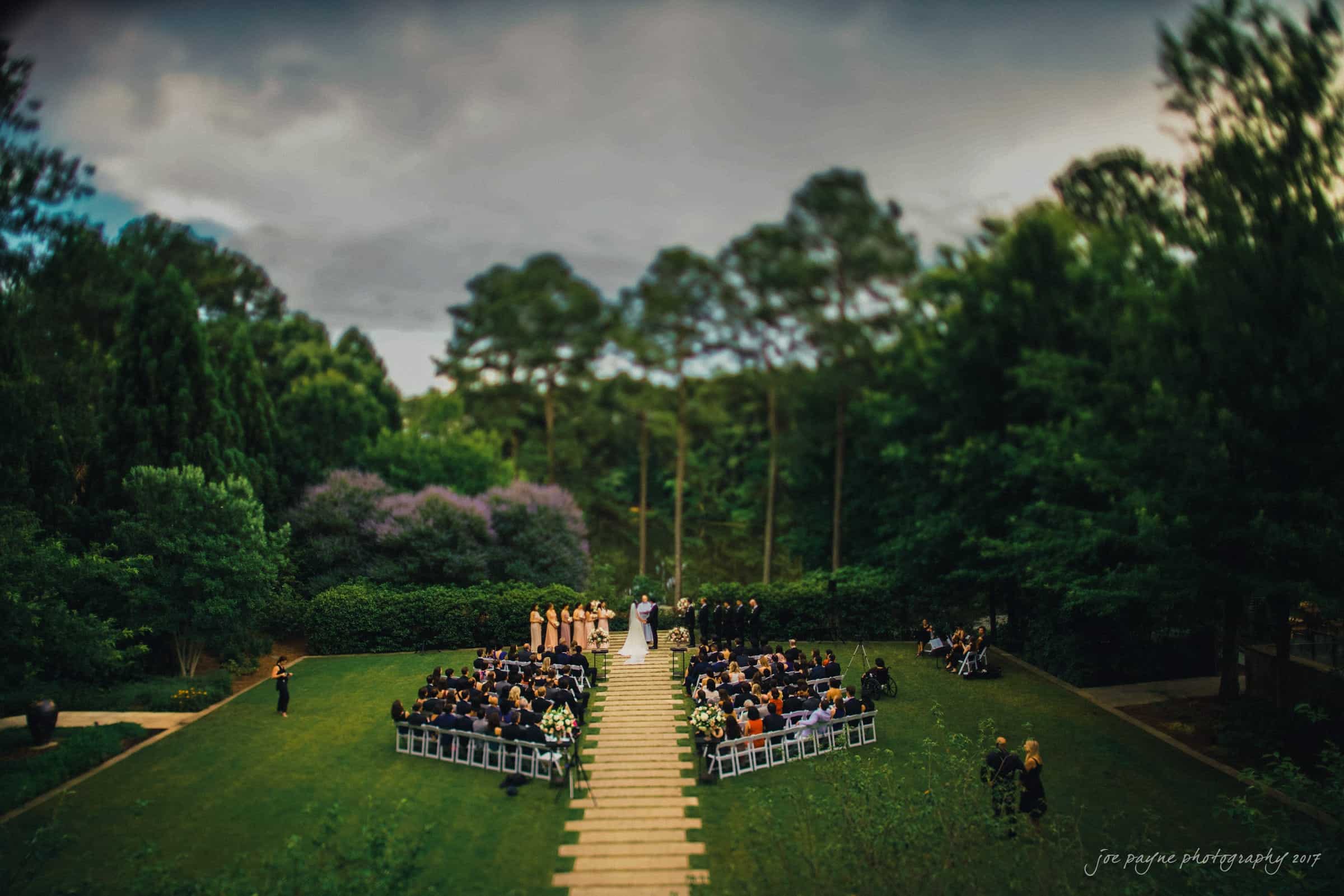 An outdoor wedding ceremony set in a lush green garden. The bride and groom stand at the altar, surrounded by the wedding party in pastel colors, and guests seated on white chairs. Tall trees and a cloudy sky serve as the backdrop.