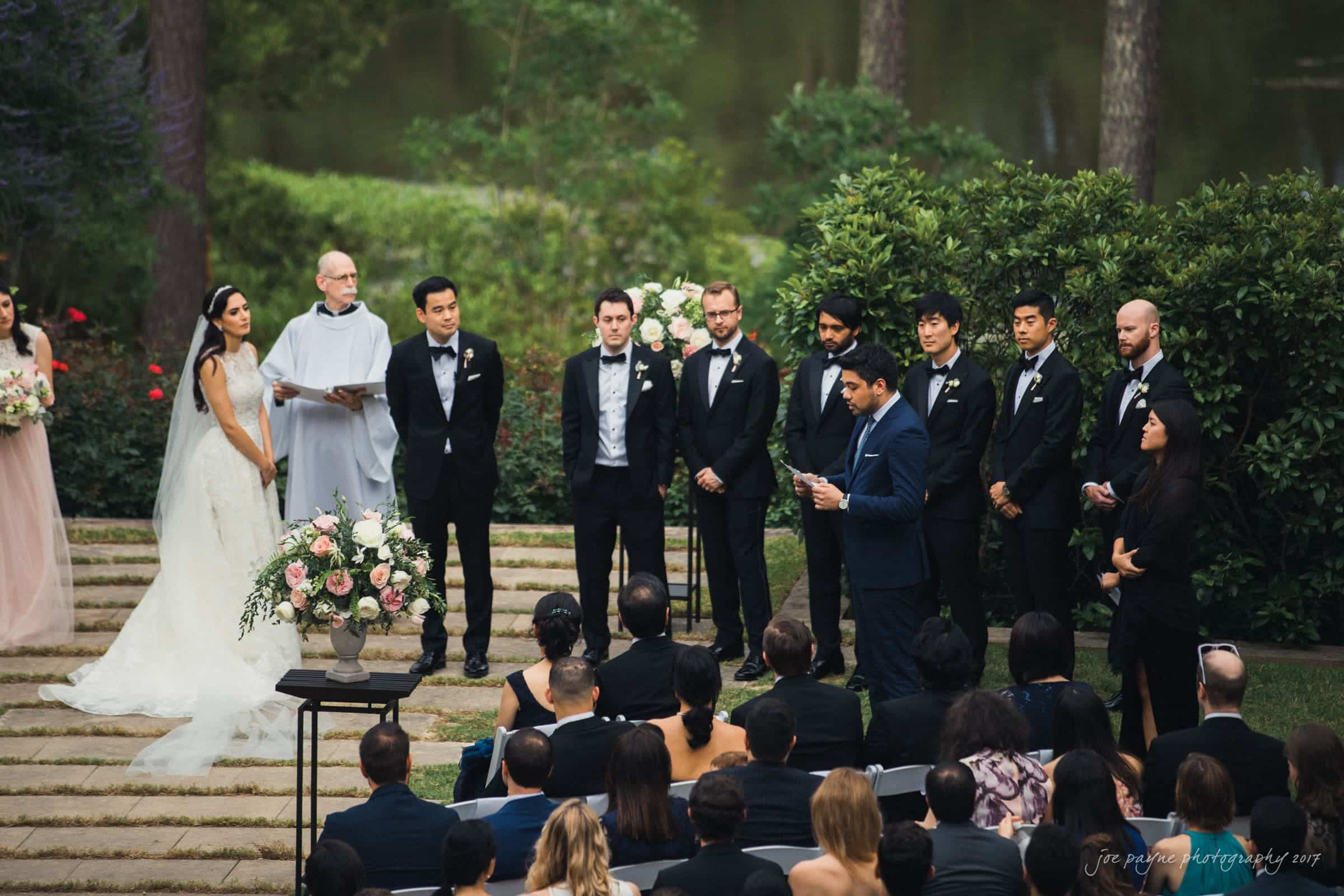 A bride and groom stand at an outdoor wedding ceremony, surrounded by groomsmen and bridesmaids in formal wear. The officiant, wearing white, is reading. Guests are seated, watching the event. Lush greenery and a pond are visible in the background.