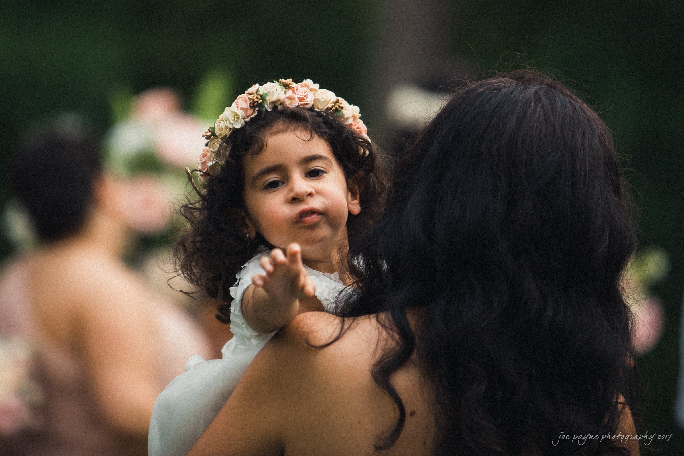 A young child with curly hair and a floral crown reaches out while being carried by an adult with long dark hair. The background is blurred, featuring soft green and pink tones.