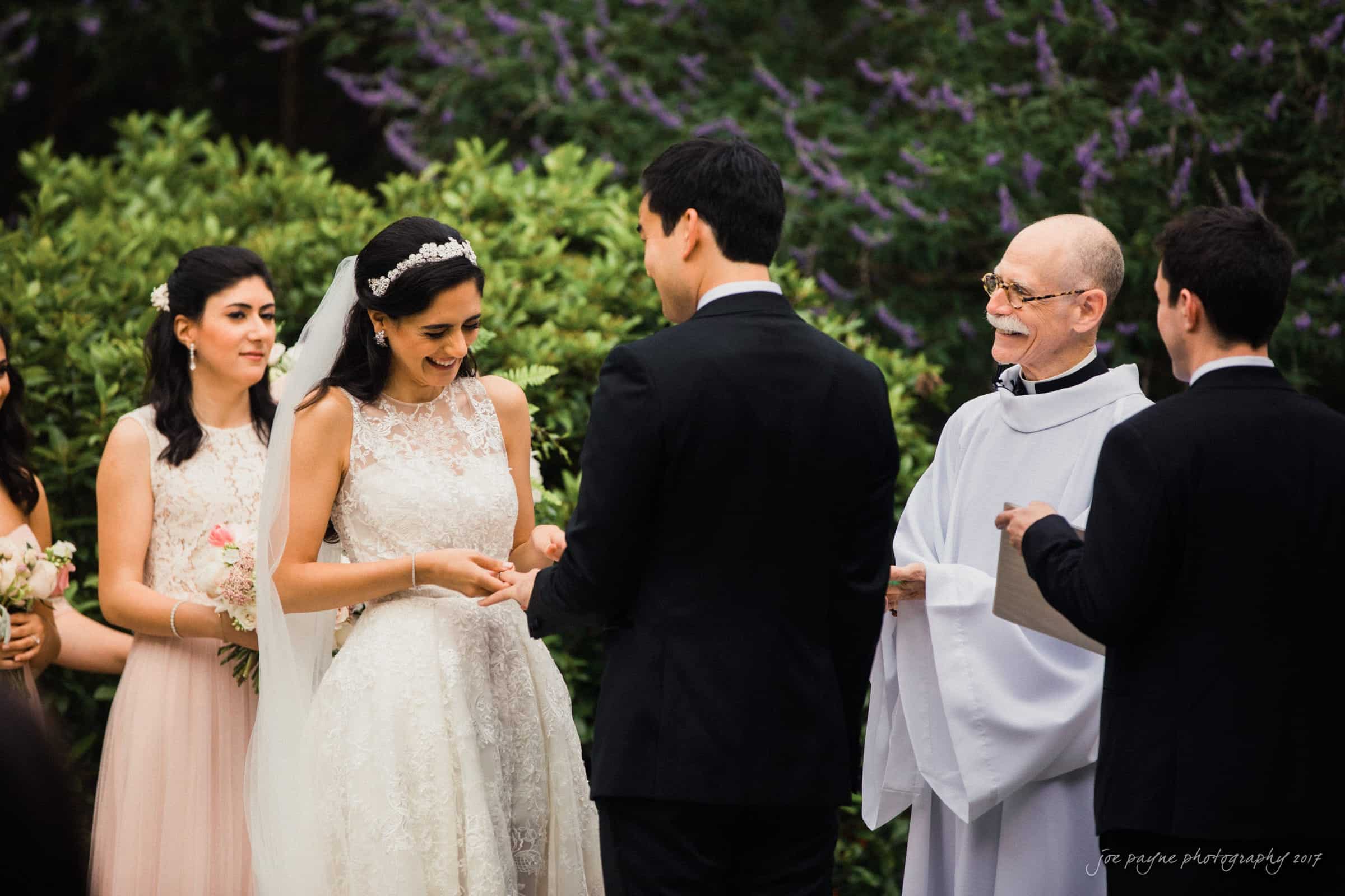 A bride in a white gown and veil exchanges rings with the groom in a black suit during an outdoor wedding ceremony. Bridesmaids stand nearby holding flowers, and a smiling officiant in white robes holds a book. Lush greenery and purple flowers form the backdrop.