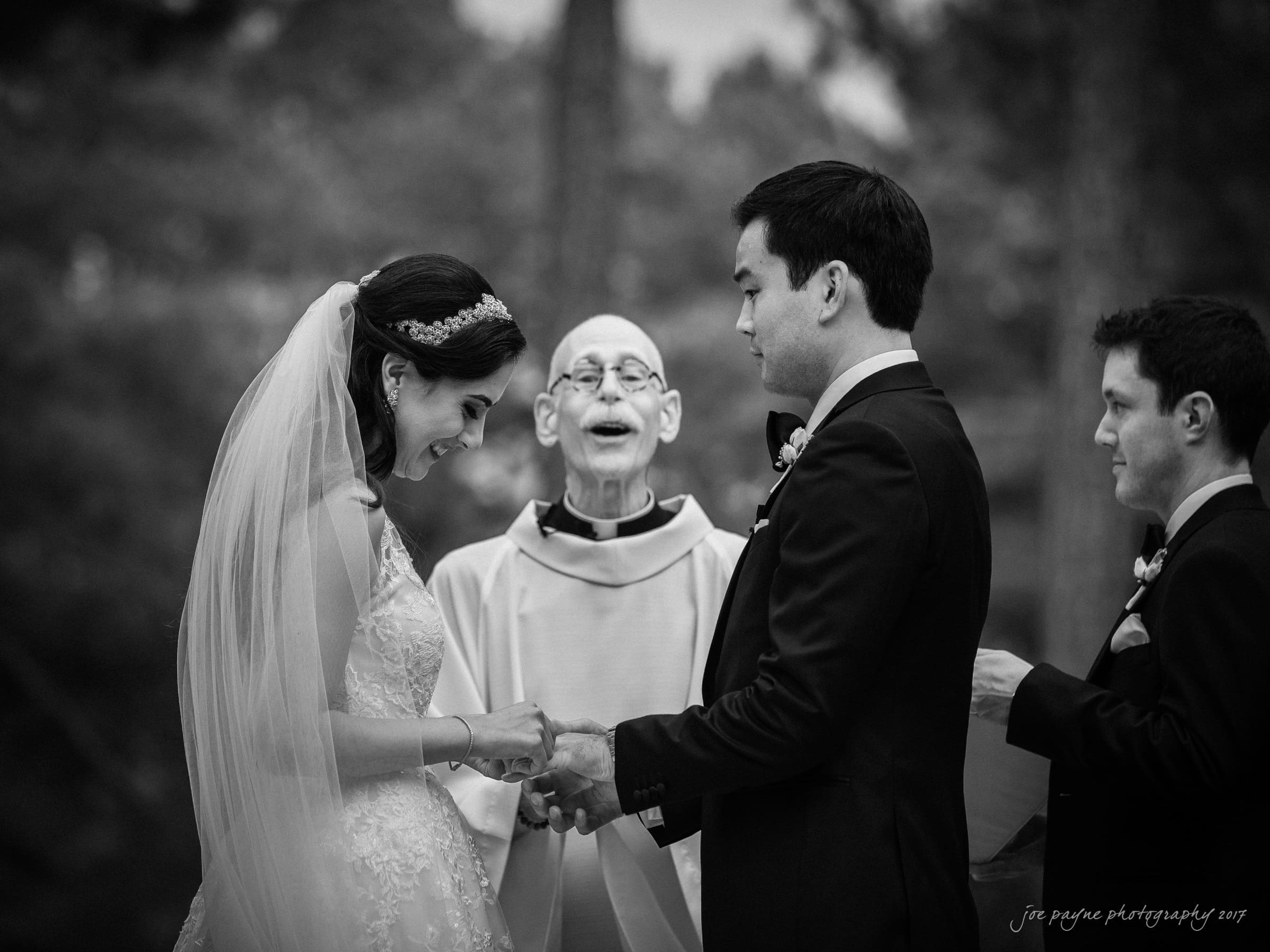 A bride and groom stand holding hands during an outdoor wedding ceremony. The bride wears a veil and a lace dress, and the groom is in a suit. A clergyman between them smiles warmly, while another man in a suit stands beside them.