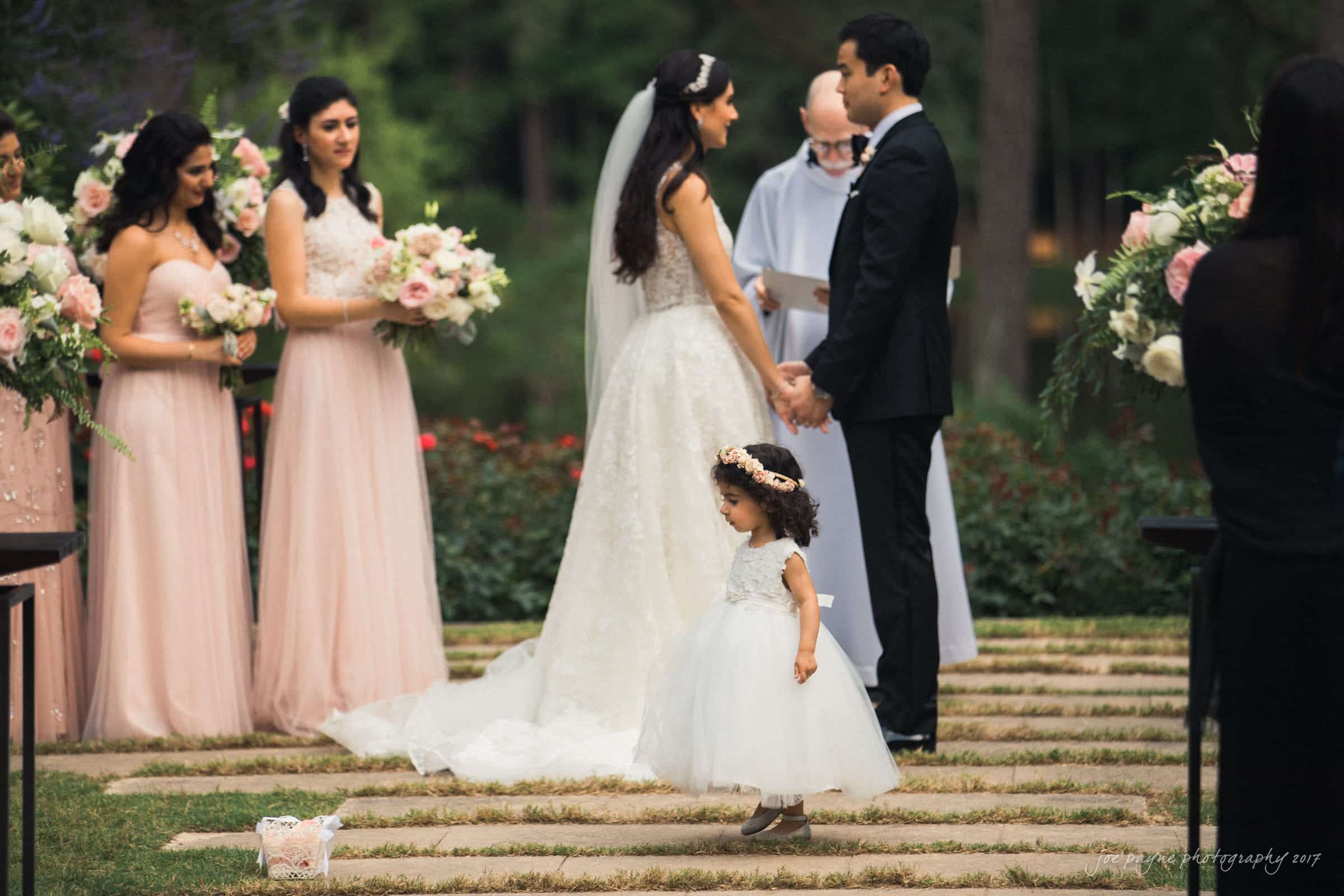 A bride and groom stand facing each other, holding hands during an outdoor wedding ceremony. Bridesmaids in pink dresses stand to the side, holding bouquets. A young flower girl in a white dress is in the foreground, looking away.