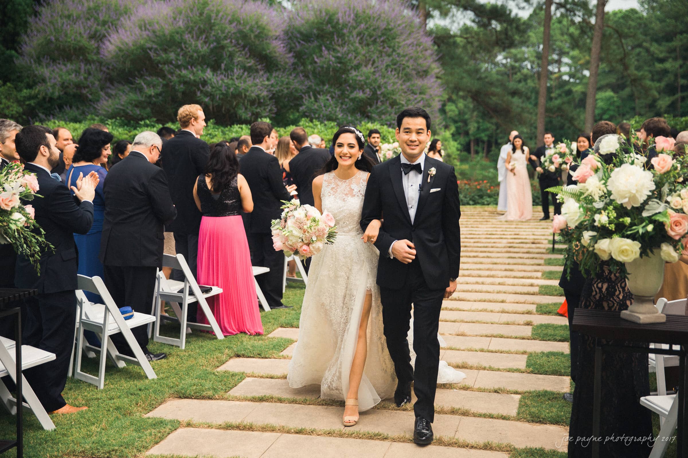 A bride and groom joyfully walk down an outdoor aisle lined with guests, holding bouquets and applauding. The garden setting features lush greenery and purple flowers, creating a romantic atmosphere. The couple is dressed elegantly in a wedding gown and tuxedo.