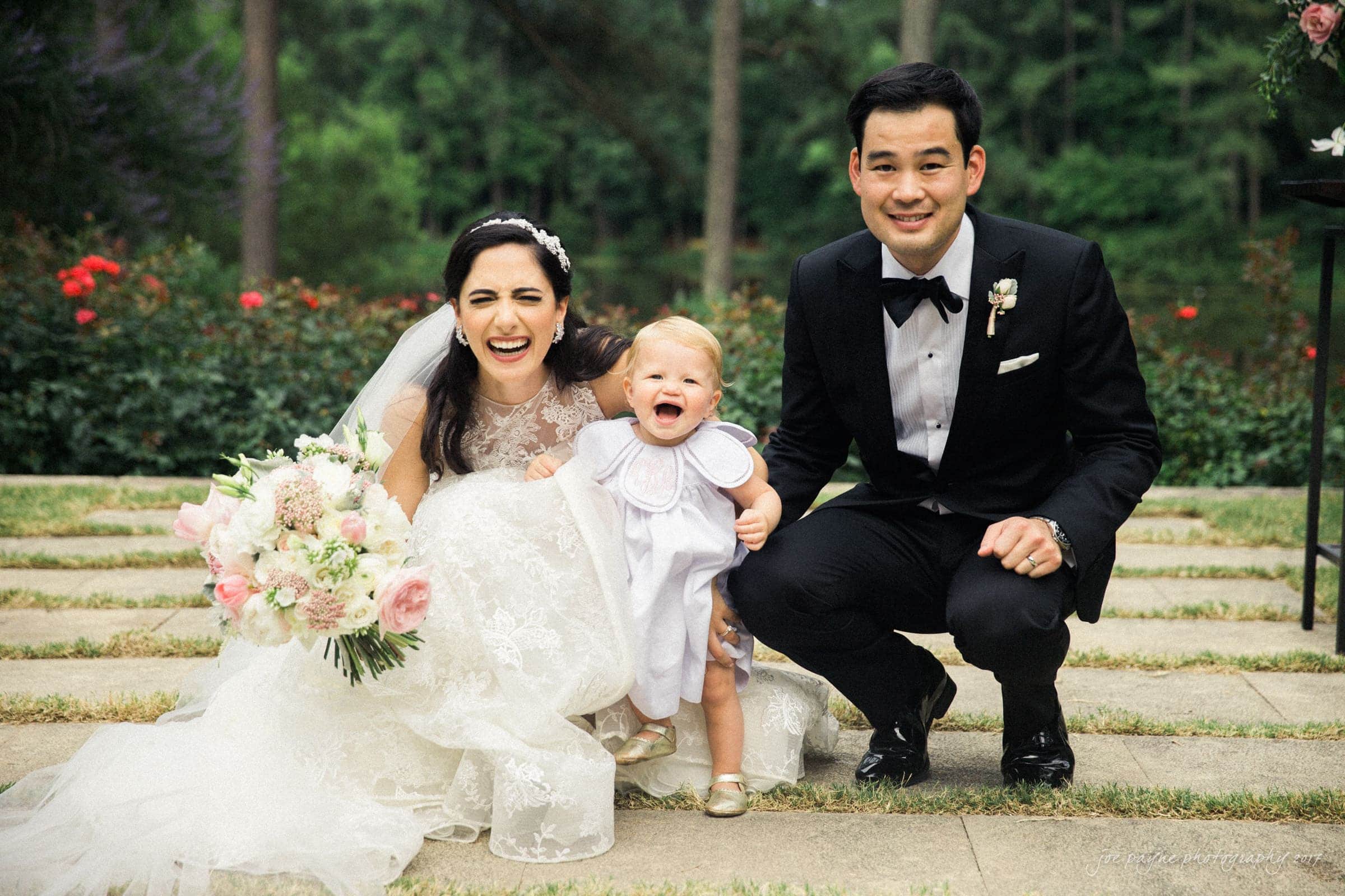 A bride and groom in formal attire smile joyfully, kneeling beside a happy toddler in a white dress. They are outdoors, surrounded by greenery and flowers, on a sunny day.
