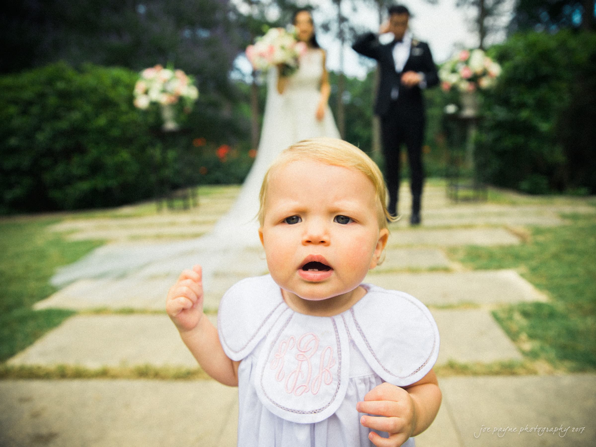 A toddler with blonde hair and a white outfit stands in the foreground of a wedding scene, looking curious. Blurred in the background, a bride and groom are smiling, surrounded by greenery and floral arrangements.