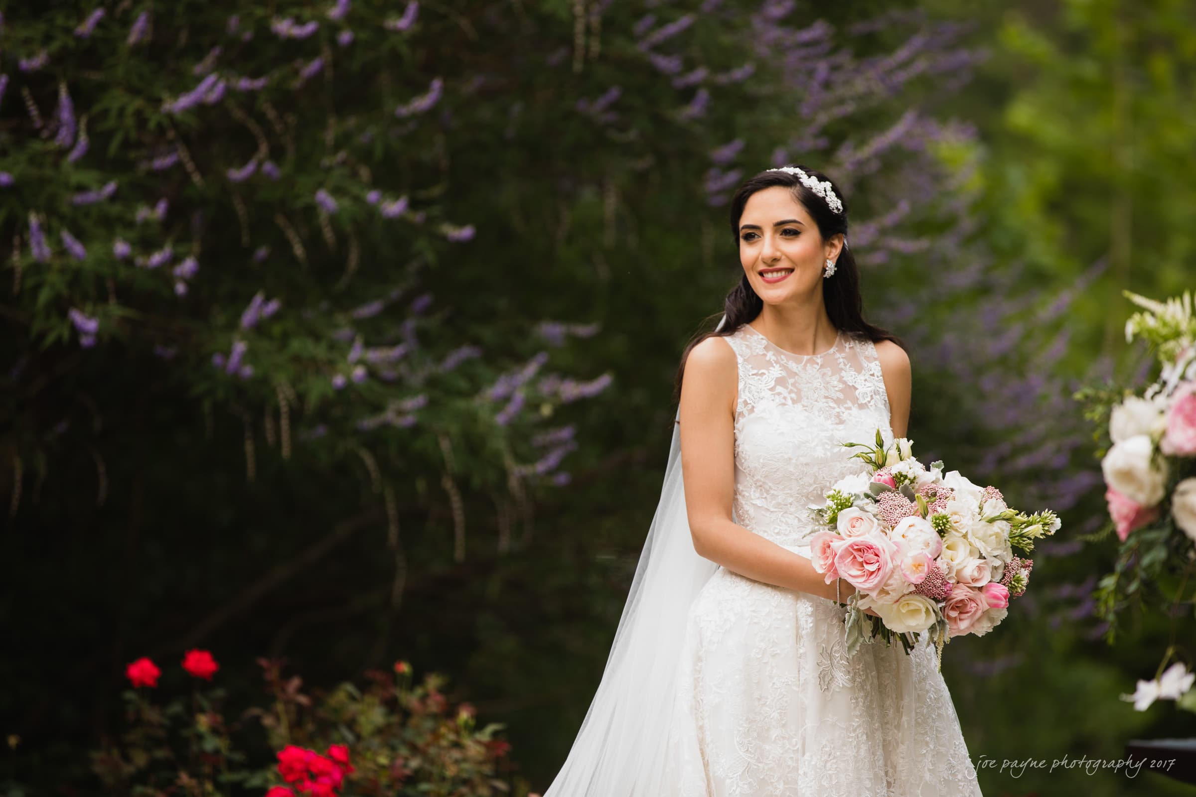A bride in a lace wedding dress holds a bouquet of pink and white flowers. She stands outdoors with lush green foliage and purple flowers in the background, smiling gently.