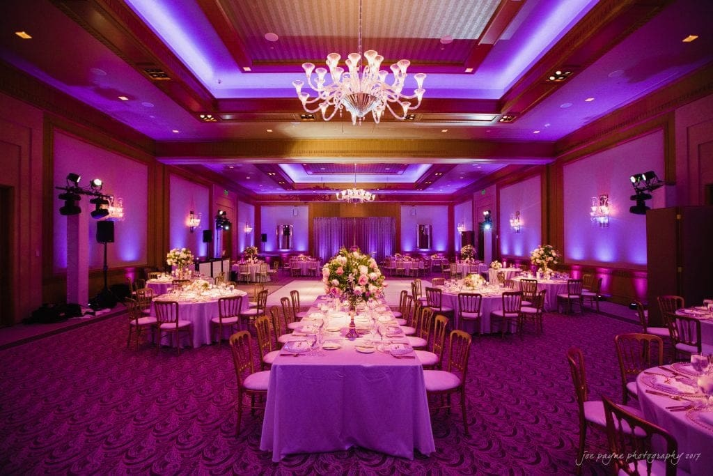 A beautifully decorated banquet hall with elegant chandeliers and purple lighting. The room is set with round and long tables adorned with white tablecloths and floral centerpieces. The ambiance is luxurious and inviting.