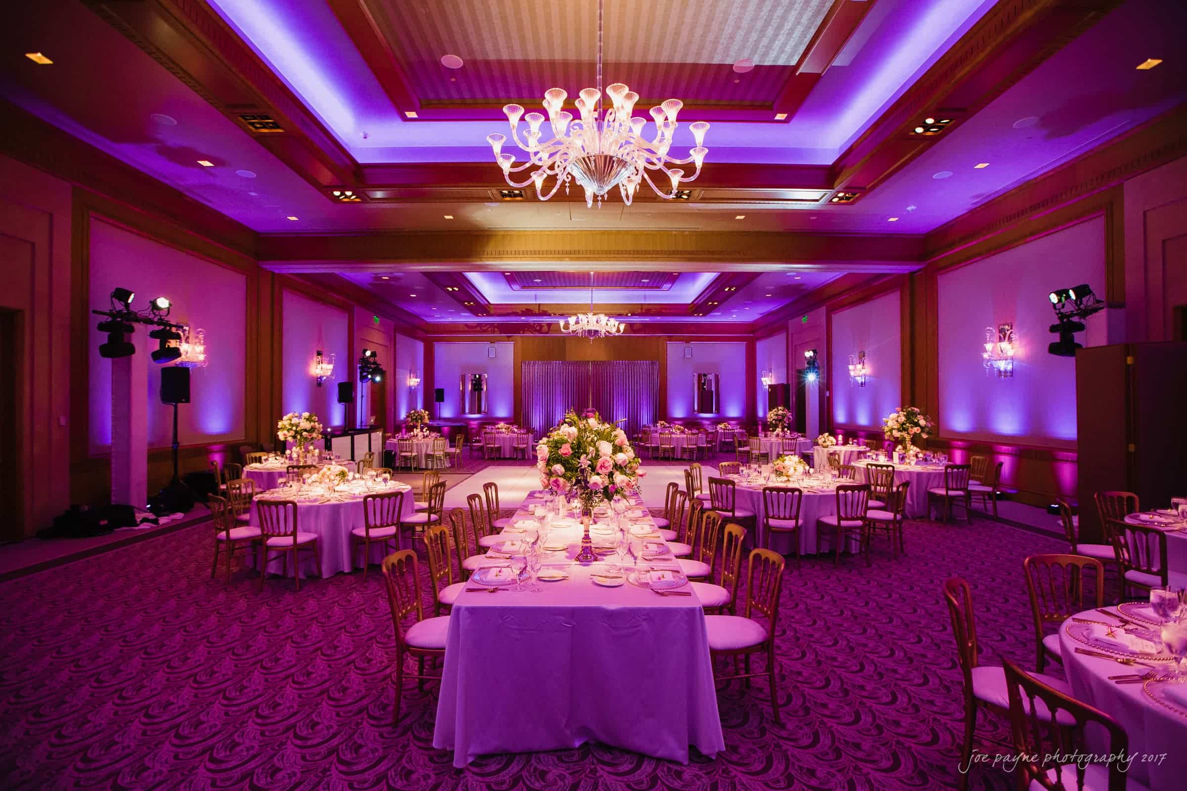 A beautifully decorated banquet hall with elegant chandeliers and purple lighting. The room is set with round and long tables adorned with white tablecloths and floral centerpieces. The ambiance is luxurious and inviting.