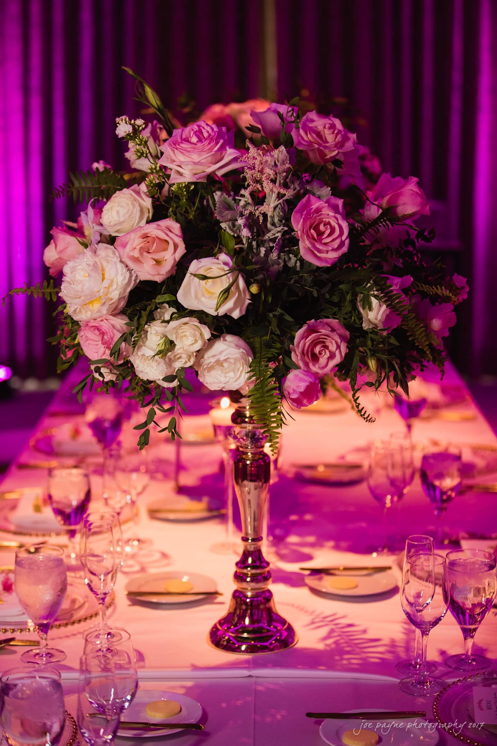 A lavish floral centerpiece with pink and white roses on a metallic stand adorns a long table set with glassware and plates, under purple ambient lighting.