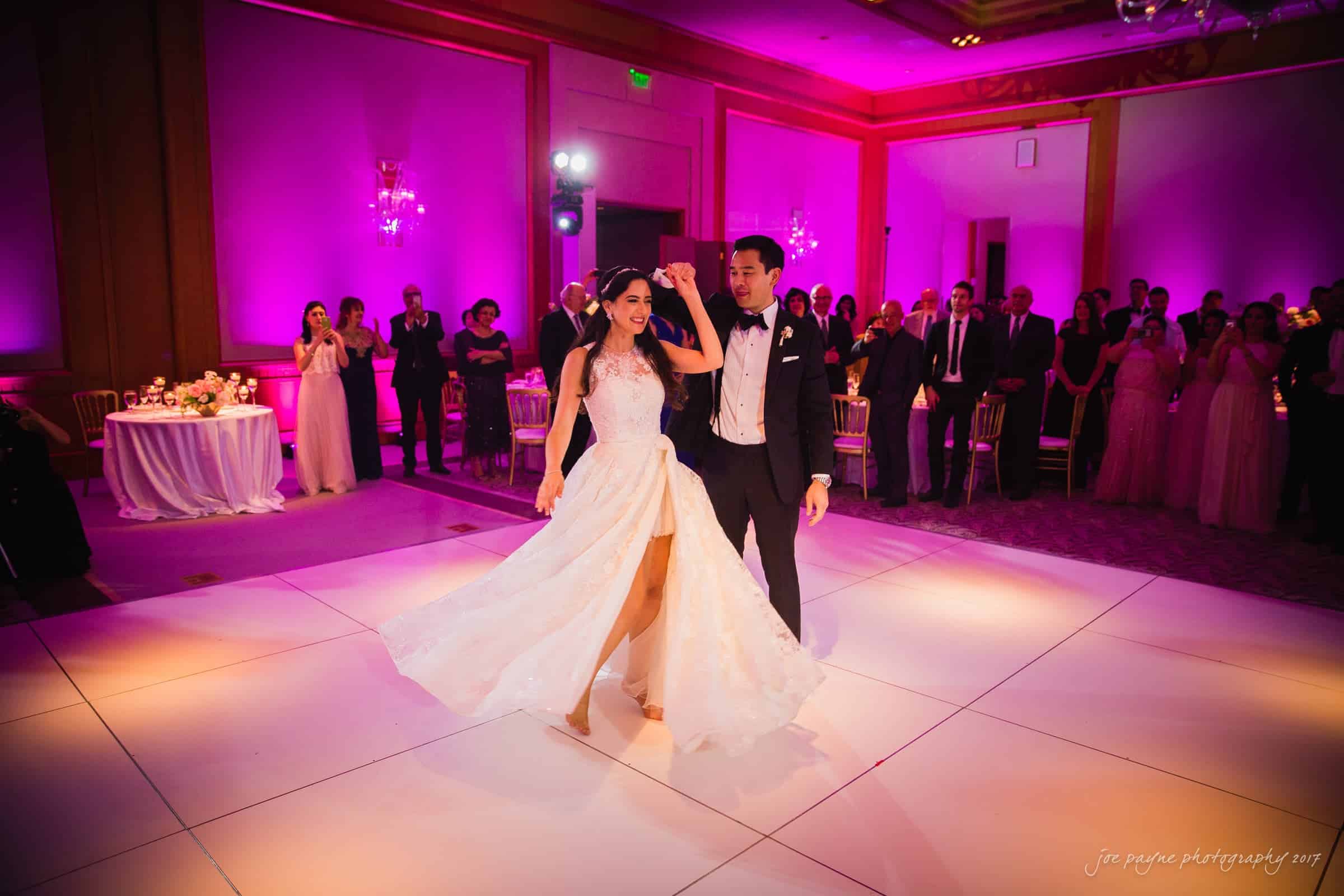 A bride and groom dance on a brightly lit dance floor at their wedding reception. The room is filled with guests watching, and the walls are illuminated with pink lighting. The brides dress flows elegantly as they twirl.