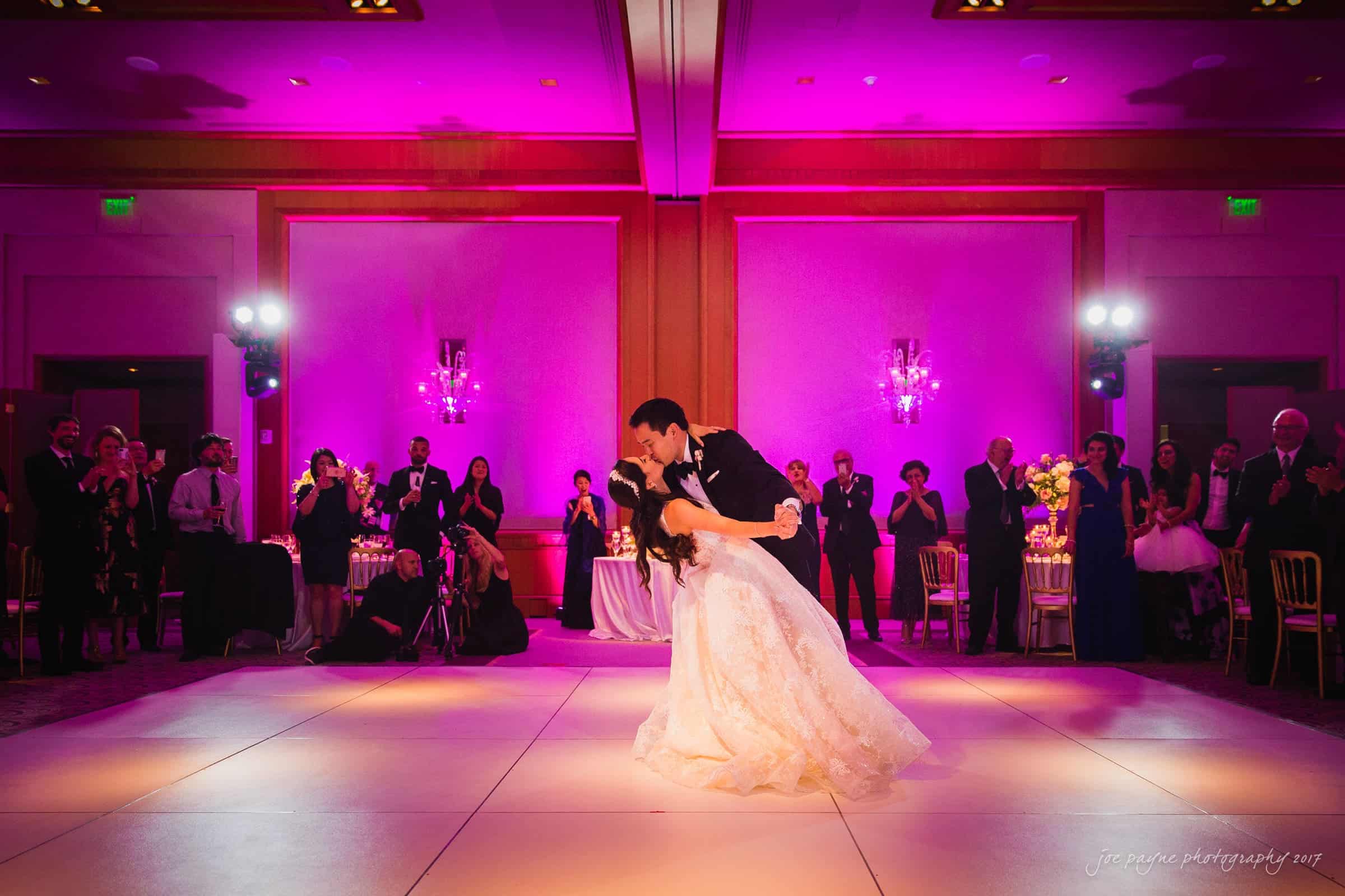 A couple shares a romantic dance under vibrant purple lighting at a wedding reception. Guests in formal attire surround the dance floor, watching and clapping. The elegant setting includes chandeliers and floral arrangements.