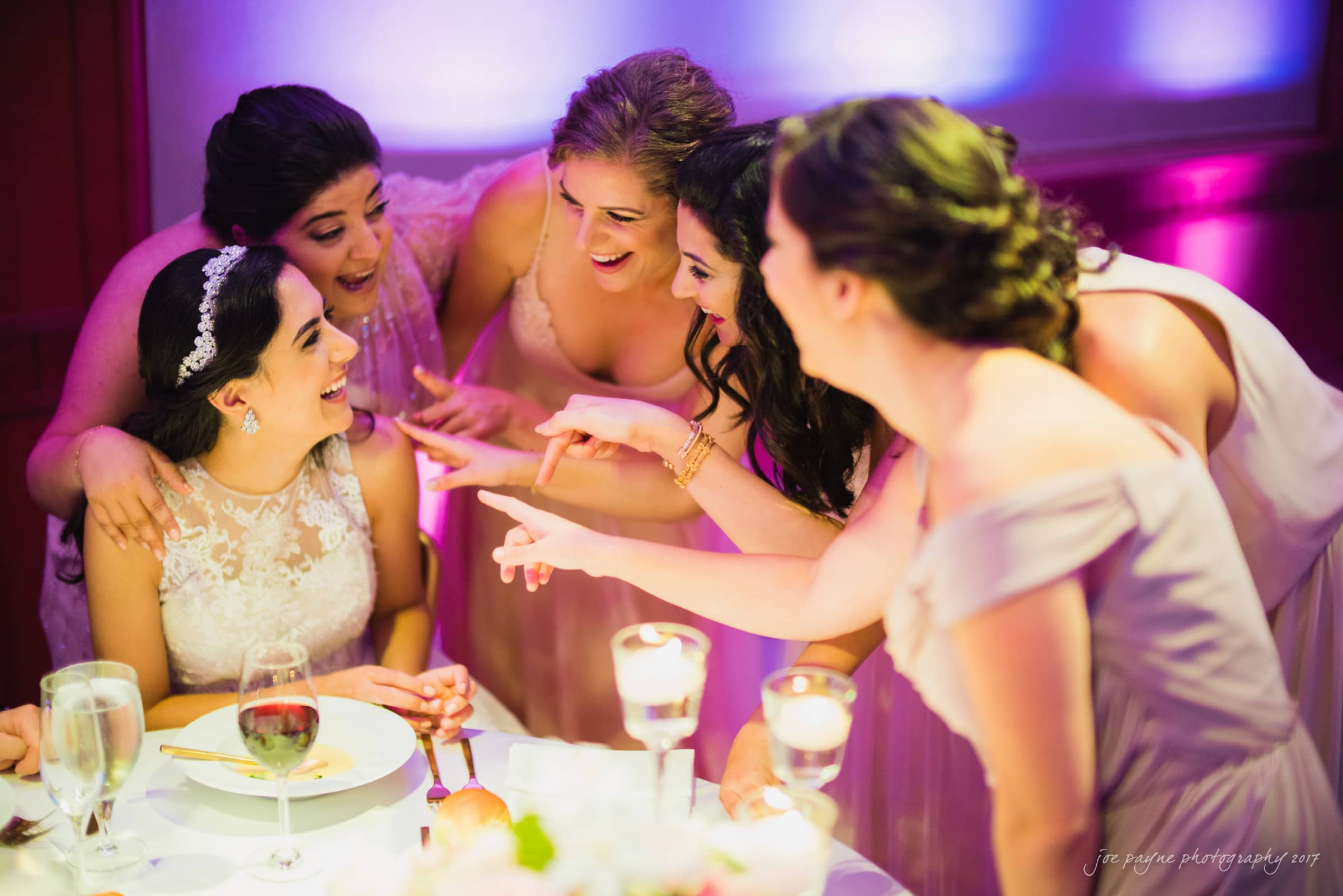 A bride sitting at a table is joyfully surrounded by five bridesmaids. They are all smiling and playfully gesturing towards her. The scene is warmly lit, and the table is set with glasses and plates.