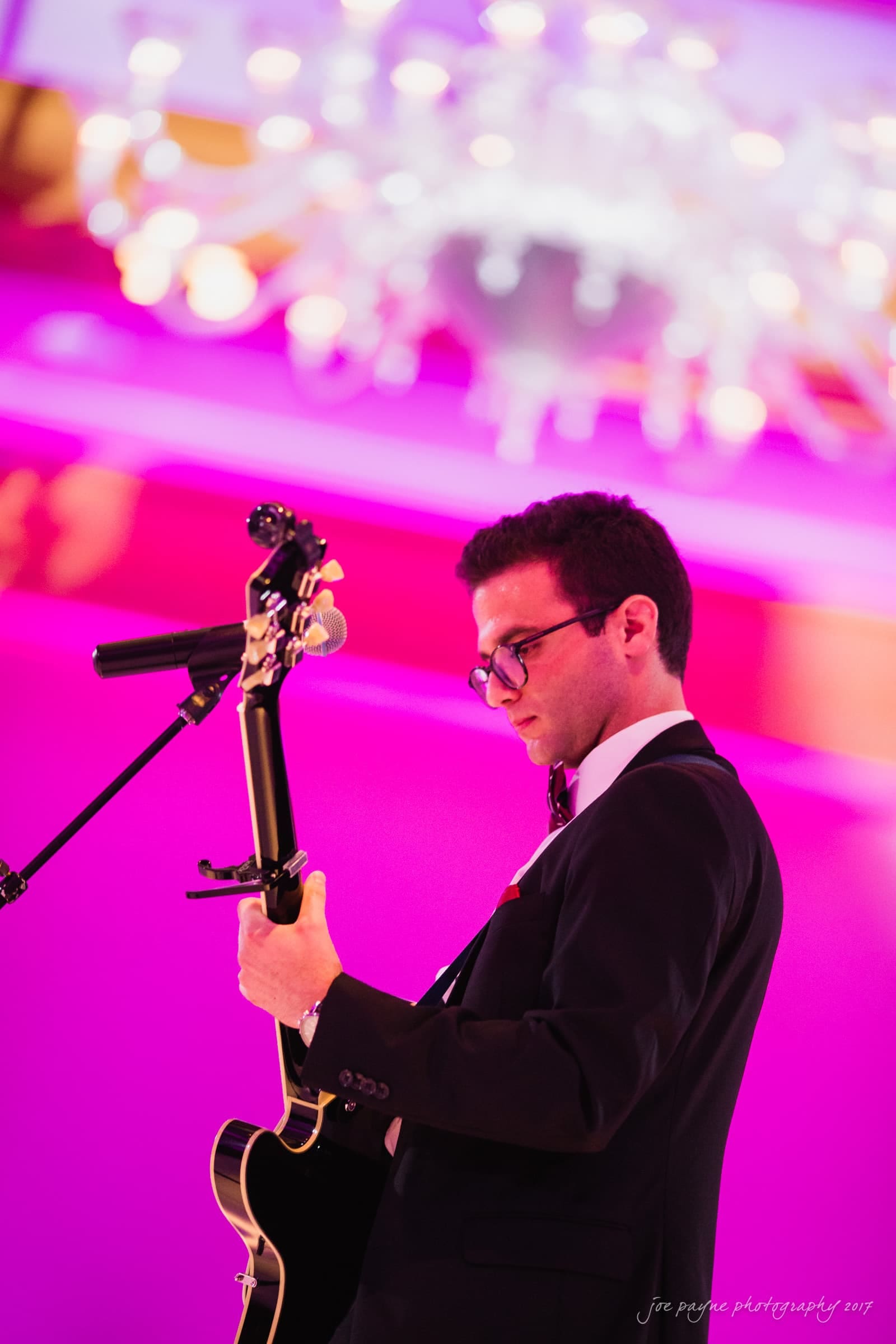 A man in a suit plays an electric guitar and sings into a microphone on stage. The background has a vibrant purple and pink hue, with out-of-focus lights overhead, creating a dramatic and colorful ambiance.