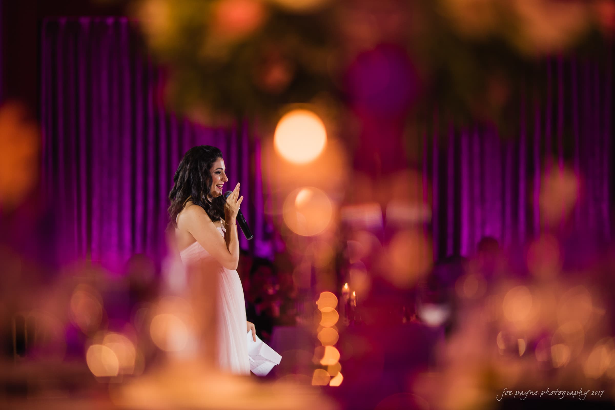 A woman in a white dress speaks with a microphone at an event decorated with purple and pink lighting. Out-of-focus flowers and lights create a soft, warm atmosphere.