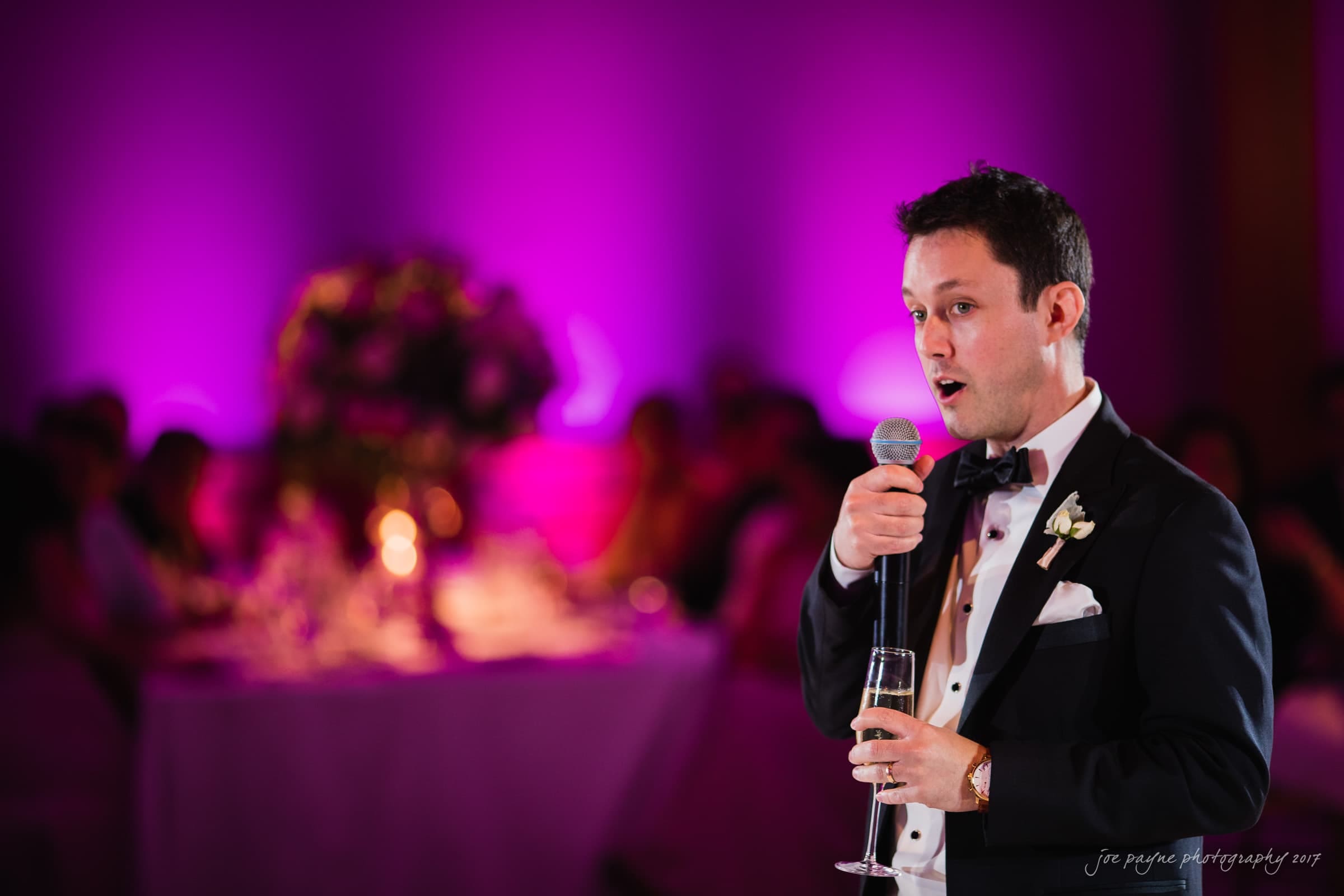 A man in a tuxedo holds a microphone and a glass, speaking at an event. The background is softly lit in purple, with a blurred view of guests seated at tables.