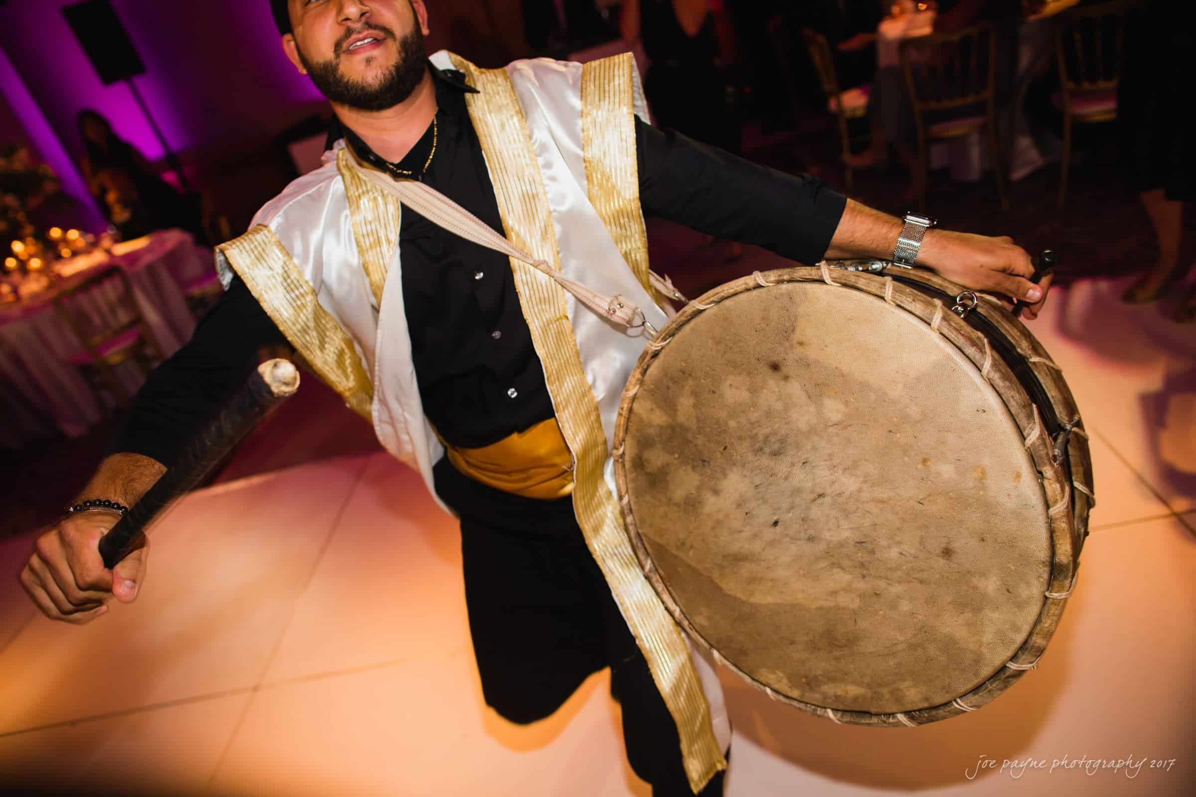 A person wearing traditional attire energetically plays a large drum at an indoor event. The background features softly lit tables and chairs, creating a festive atmosphere.