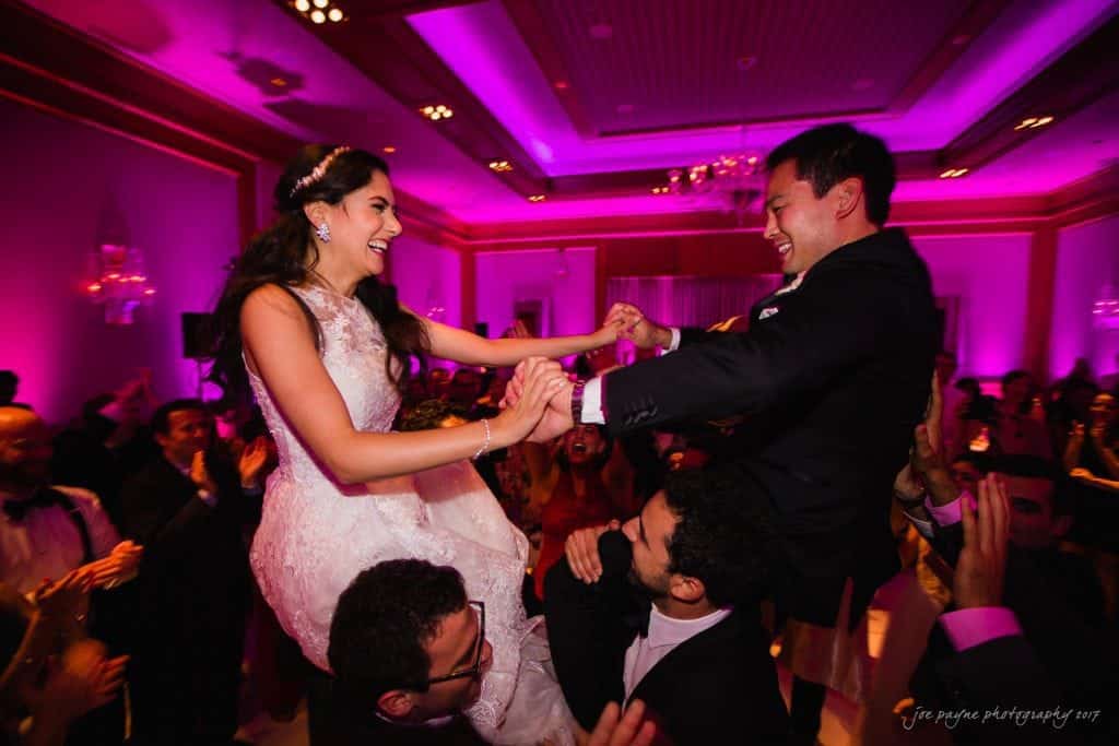 A bride and groom are joyfully lifted on chairs during a celebratory moment at a wedding reception. The room is adorned with purple lighting and surrounded by smiling guests dancing and clapping along.