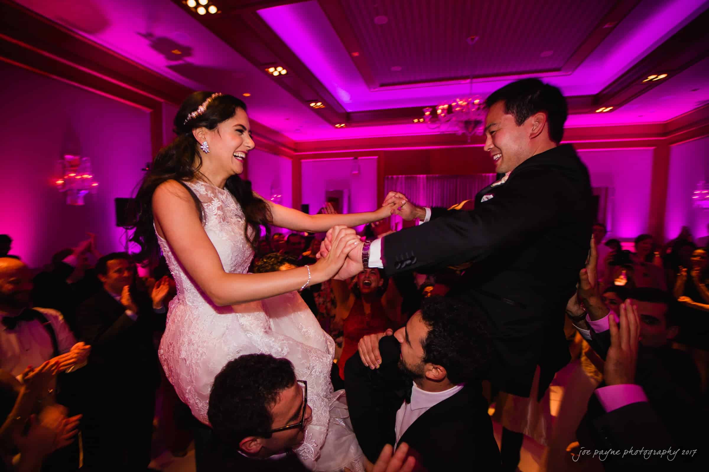 A bride and groom are joyfully lifted on chairs during a celebratory moment at a wedding reception. The room is adorned with purple lighting and surrounded by smiling guests dancing and clapping along.