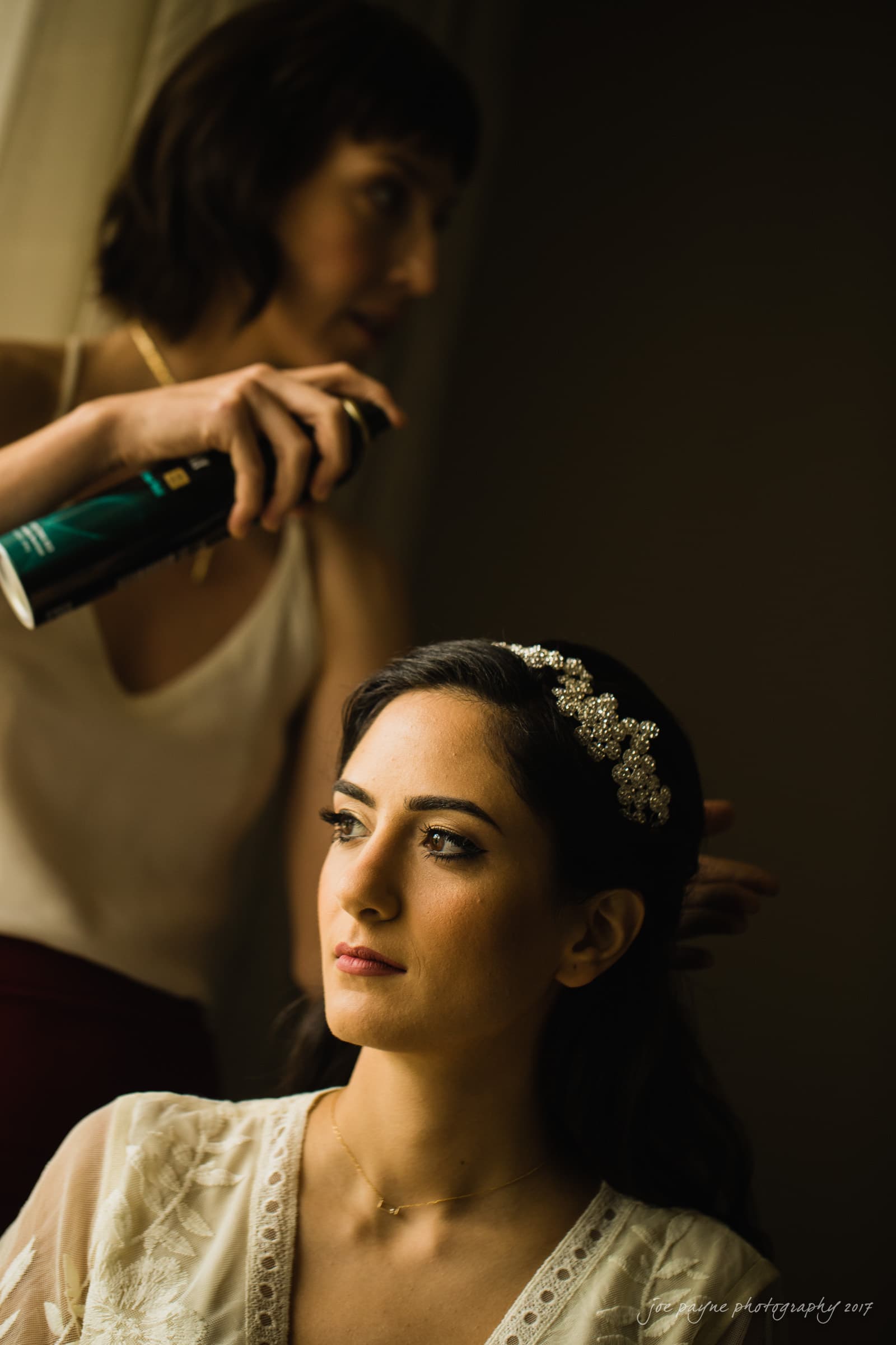 A bride with a decorative hairpiece getting her hair styled by a hairstylist who is applying hairspray. The setting appears to be a serene and softly lit room, enhancing the calm atmosphere.