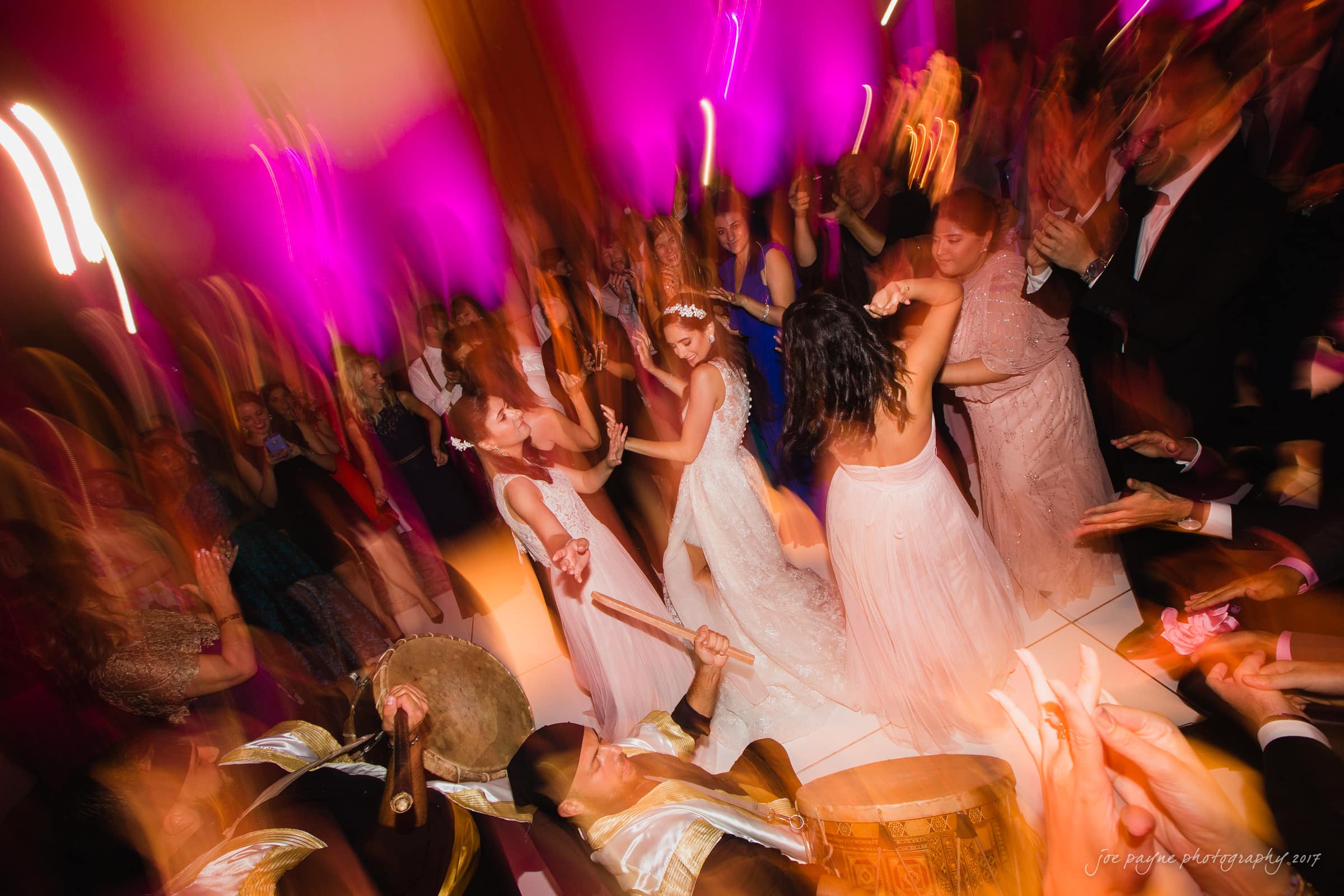 A lively wedding reception with guests dancing energetically under vibrant pink lighting. Two brides in white dresses are at the center, surrounded by joyful guests. A musician plays a drum in the foreground. The atmosphere is festive and colorful.