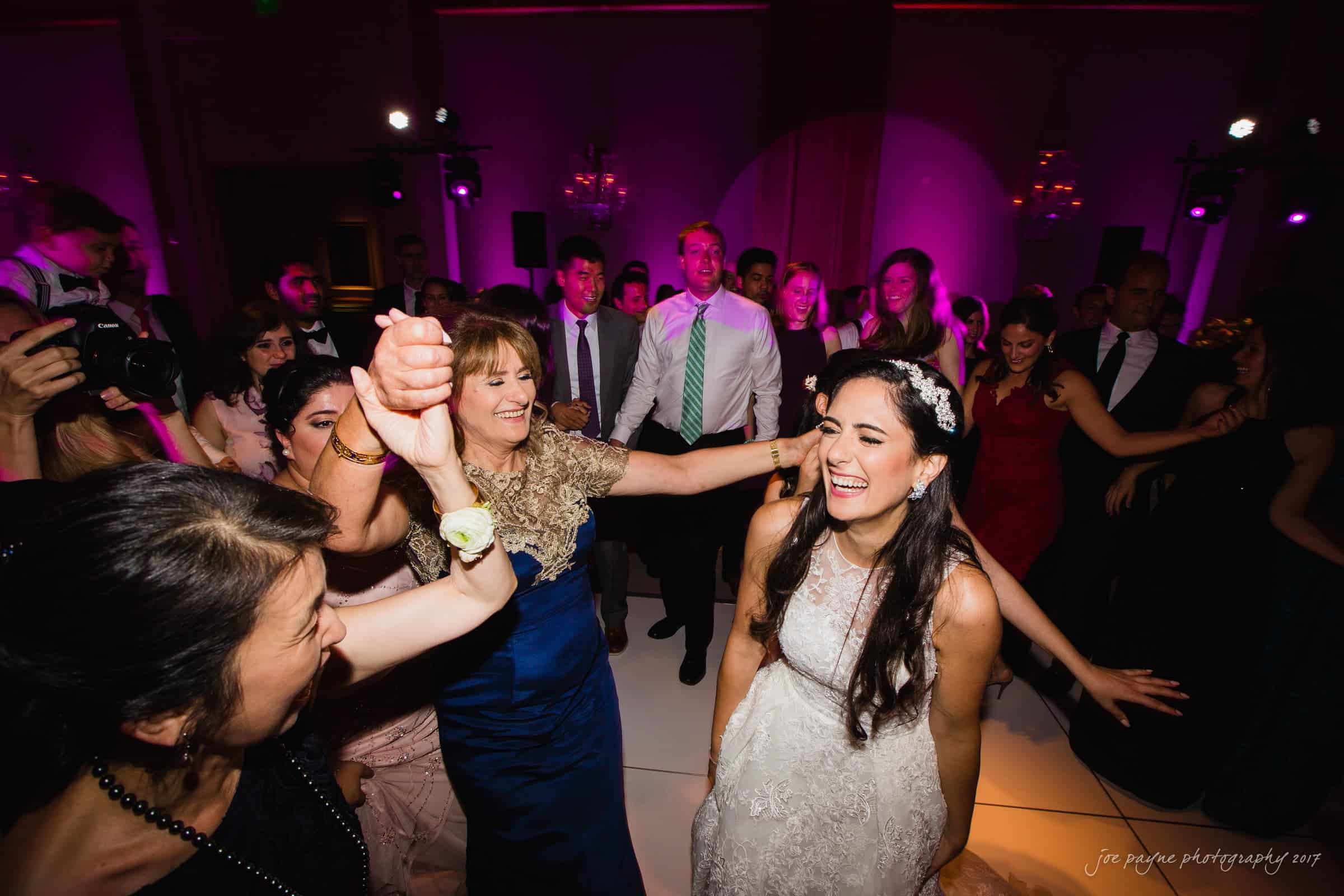 A joyful bride and guests dance in a lively, colorful setting with bright purple lighting. The bride smiles widely, surrounded by friends and family, celebrating and holding hands on the dance floor.