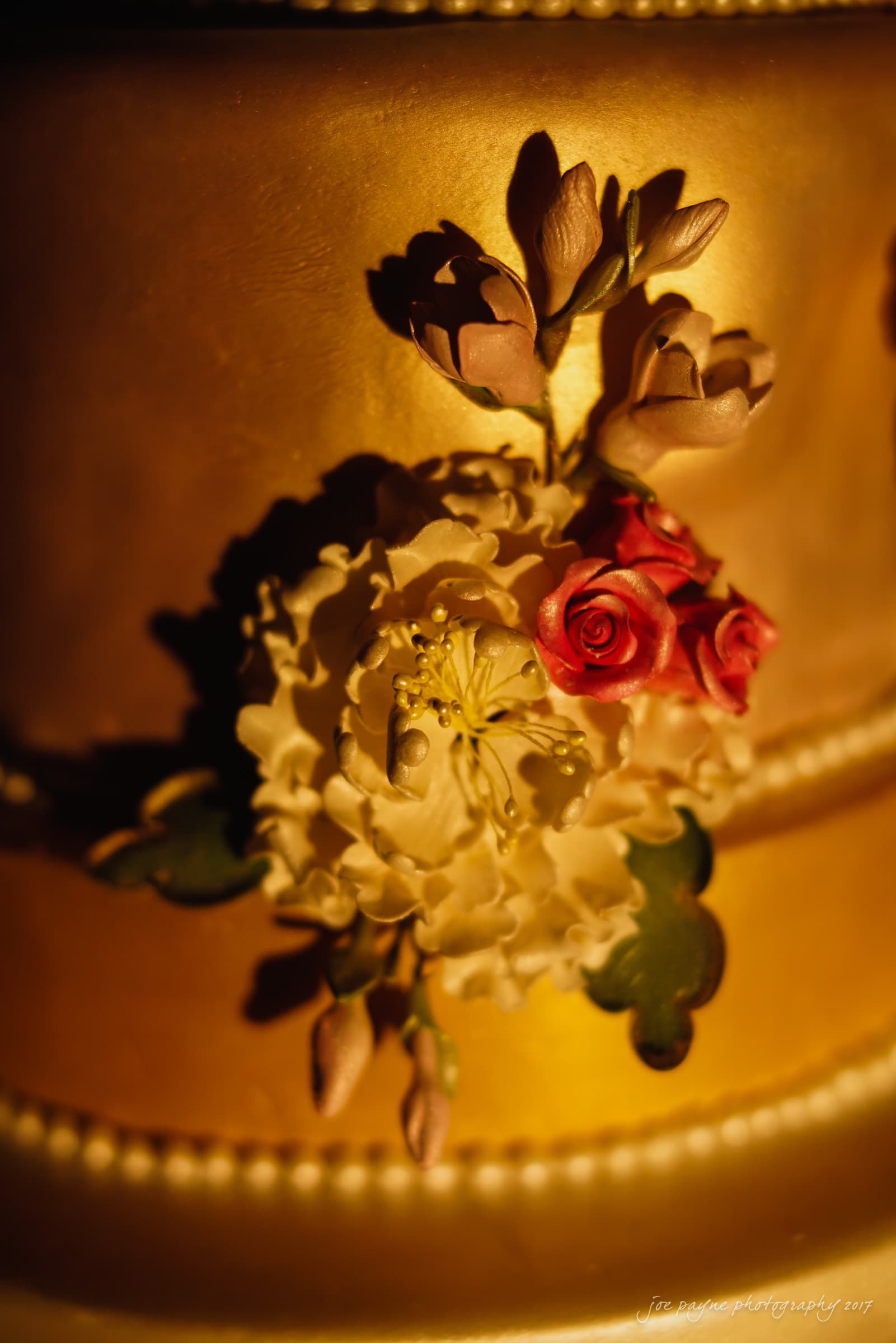 A close-up of a golden cake decorated with intricate floral designs, featuring white and red sugar flowers with green leaves, illuminated by warm lighting.