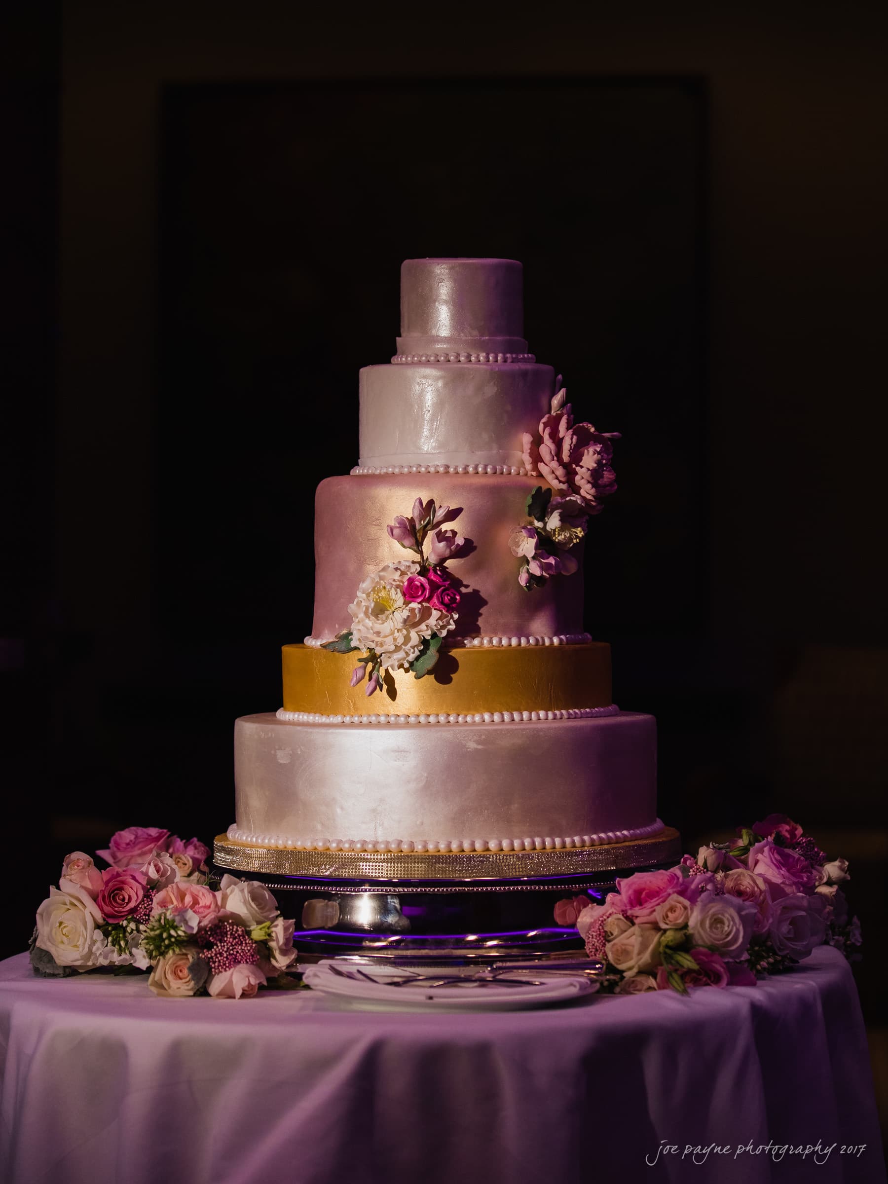 A five-tiered wedding cake with a smooth, pearlescent finish sits on a table. It is adorned with pink and white floral arrangements. The table is surrounded by additional flowers, and softly lit, creating a romantic ambiance.