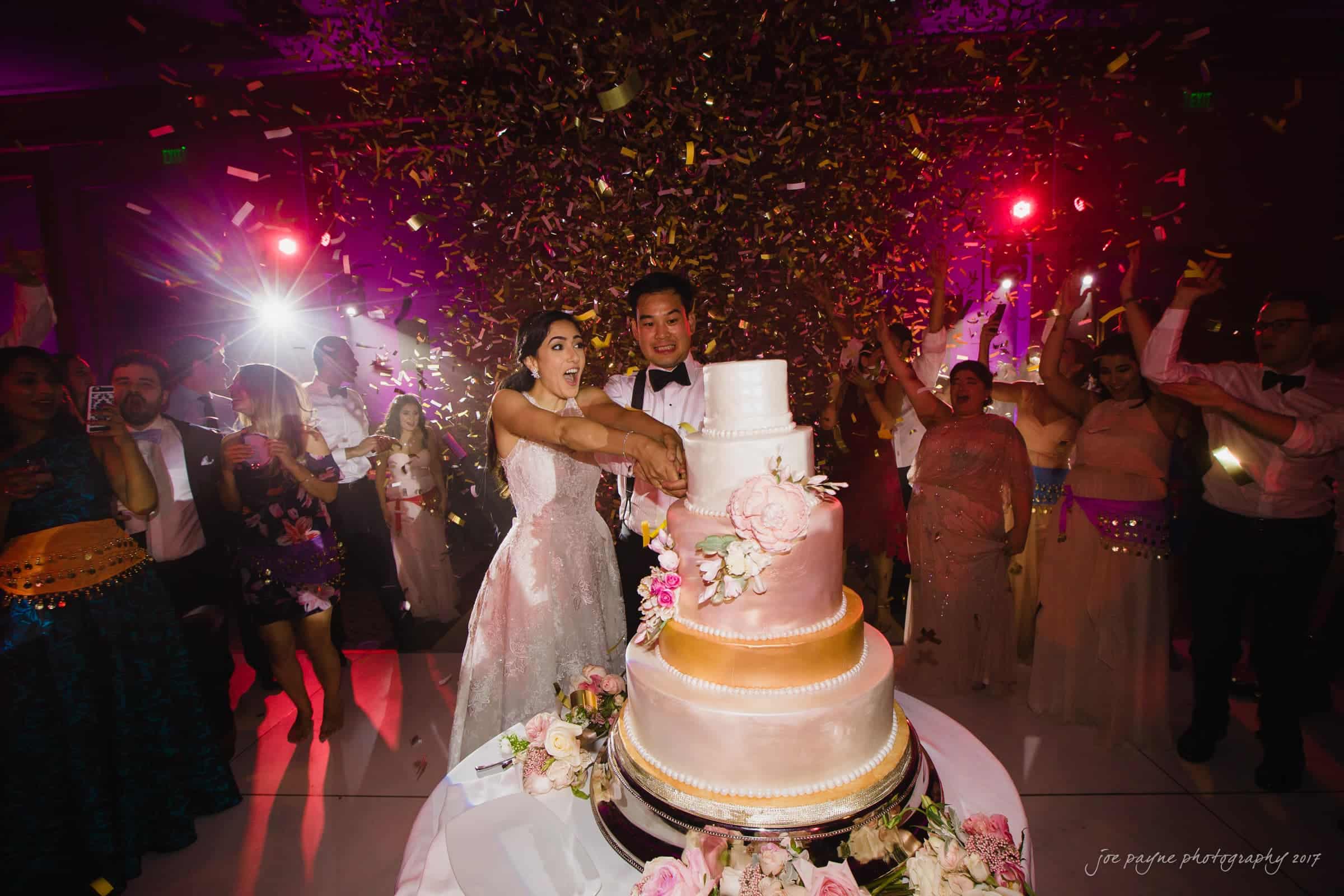 A bride and groom cut a three-tiered wedding cake surrounded by guests. Confetti fills the air, and colorful lights illuminate the scene, creating a festive atmosphere.