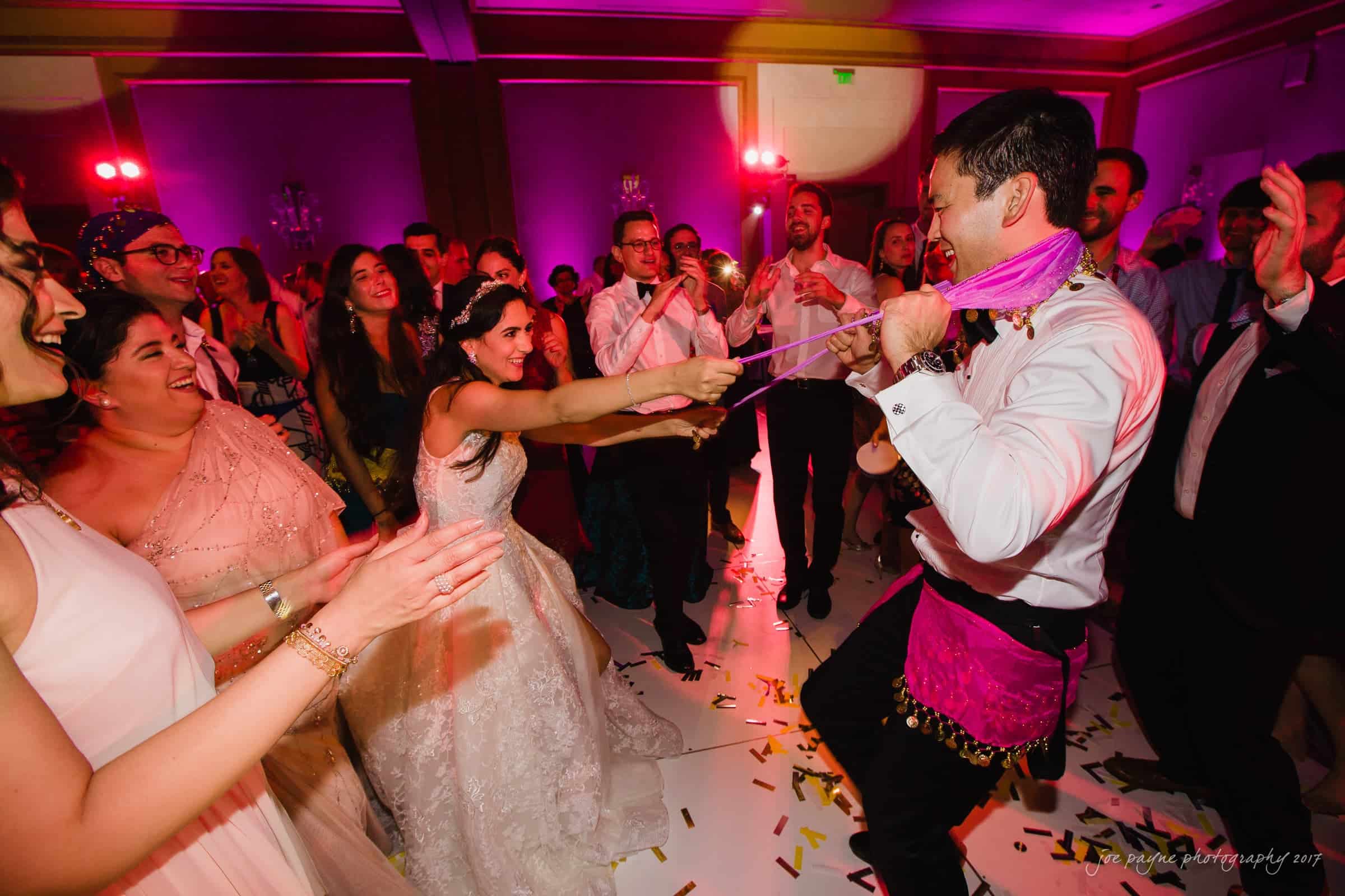 A joyful bride playfully tugs at a pink tie around the grooms neck on a dance floor. The couple is surrounded by smiling guests clapping and cheering under vibrant purple and pink lighting, with confetti scattered on the floor.