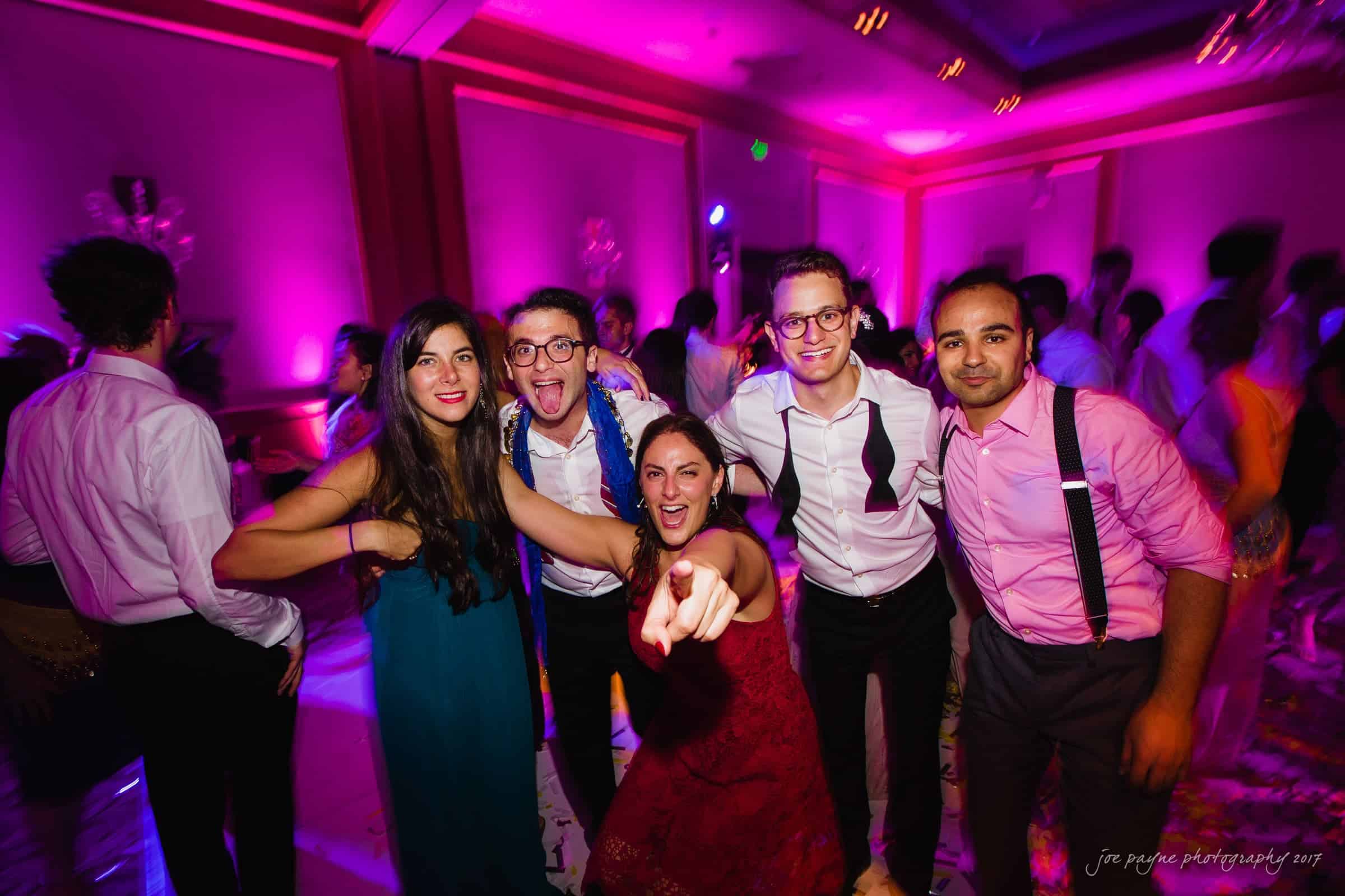 A group of five people, dressed in formal attire, pose energetically at a party with vibrant purple lighting. One person in a red dress is pointing at the camera, while the others smile and laugh around them.