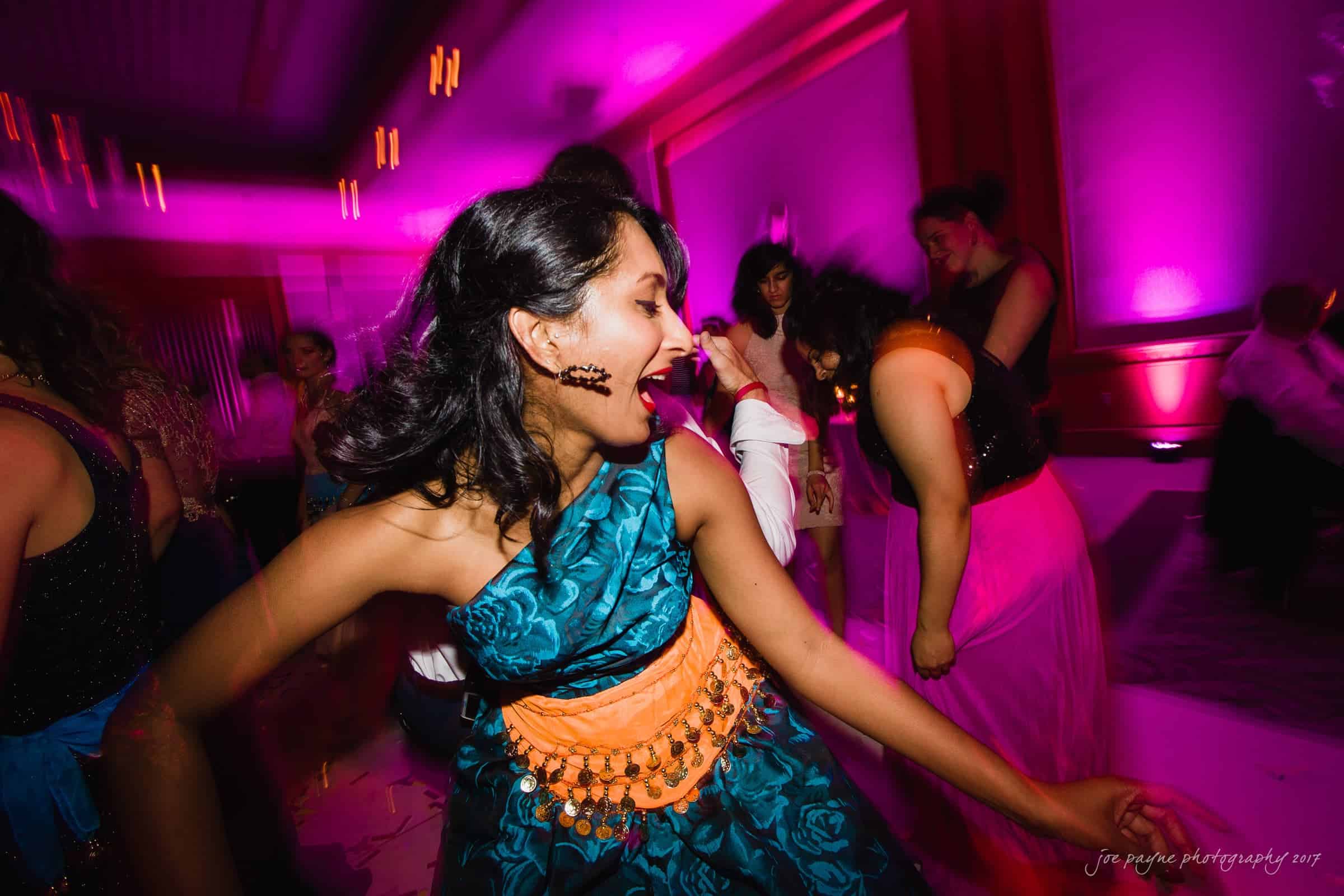 A woman in a patterned blue and orange dress joyfully dances in a vibrant, pink-lit room. Other partygoers are seen enjoying themselves in the background.