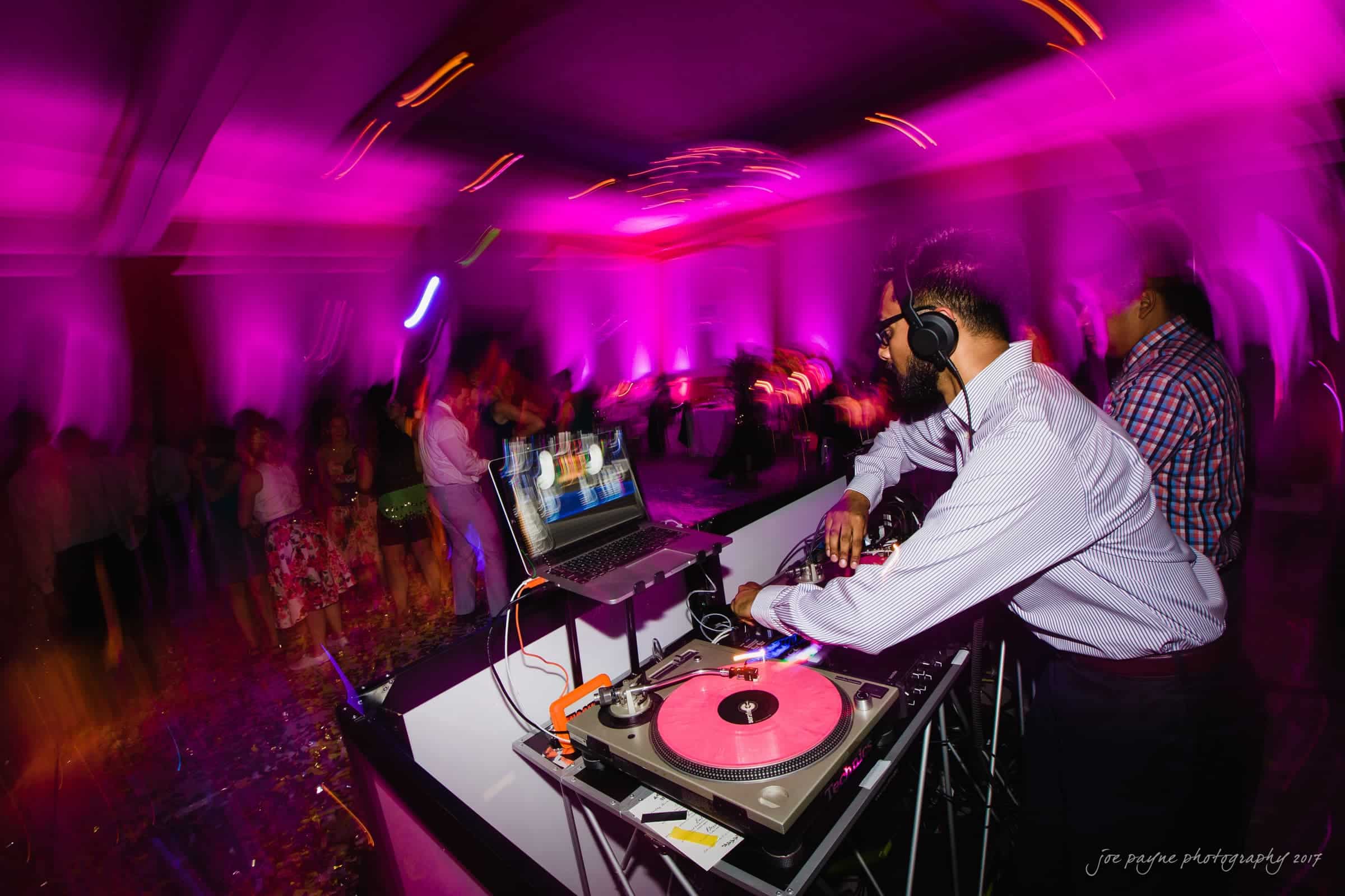 A DJ is playing music at a lively party. The room is illuminated with vibrant pink and purple lights. People are dancing in the background, and a laptop with DJ equipment and a pink vinyl record is set up on the table.