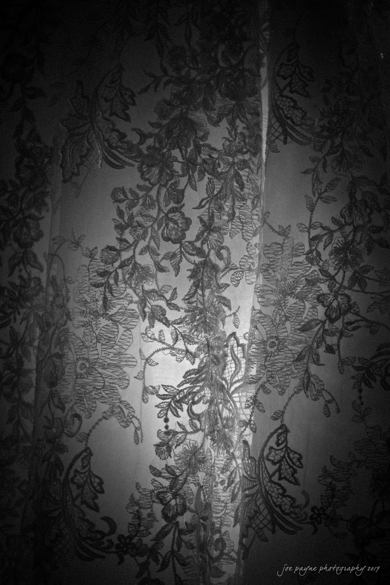 Black and white photo of a sheer lace curtain with intricate floral patterns. The backlighting creates a soft, textured effect, highlighting the delicate design.