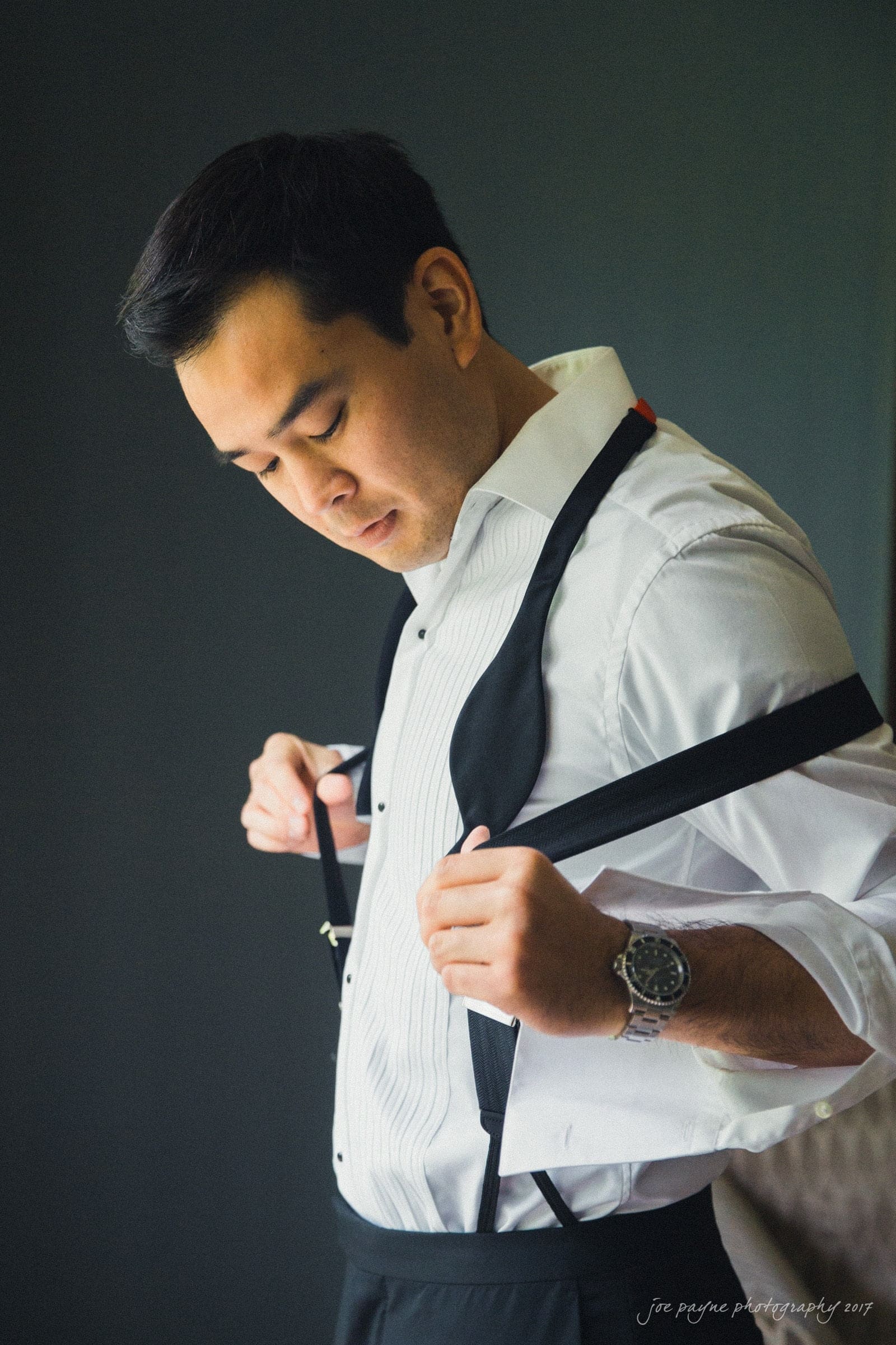 A man in formal attire adjusts his suspenders. He is wearing a white dress shirt and dark trousers, with a black vest hanging by his side. The background is dark, emphasizing his focused expression.