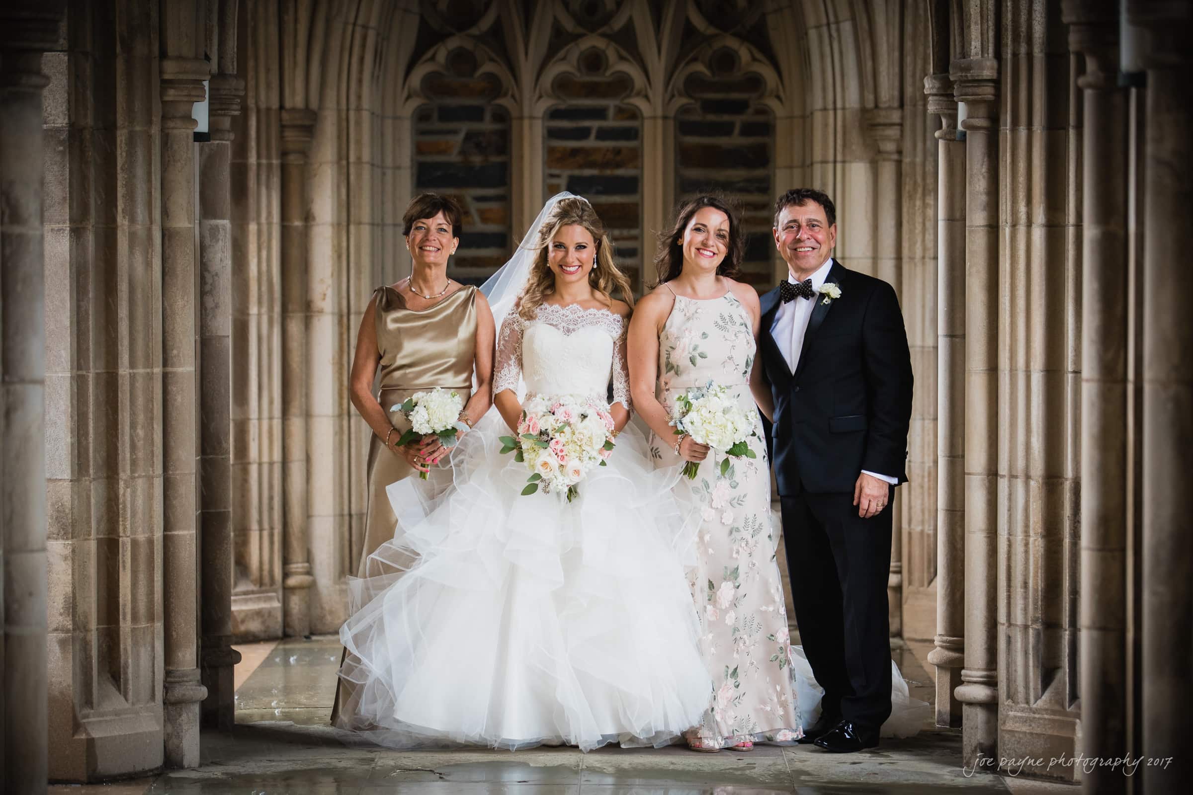 Duke Chapel Weddings A joyful wedding family portrait outside in the wet stone arcades of Duke Chapel after a rain shower, with the bride and her family all smiling happily together.
