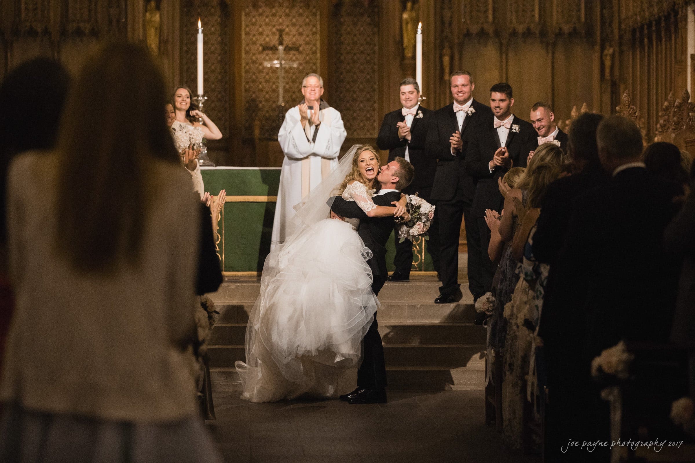 A newlywed couple shares a joyful kiss at the altar of a church, surrounded by their bridal party and guests. The bride holds her bouquet, and the groom lifts her slightly. The officiant and bridal party smile in the background.