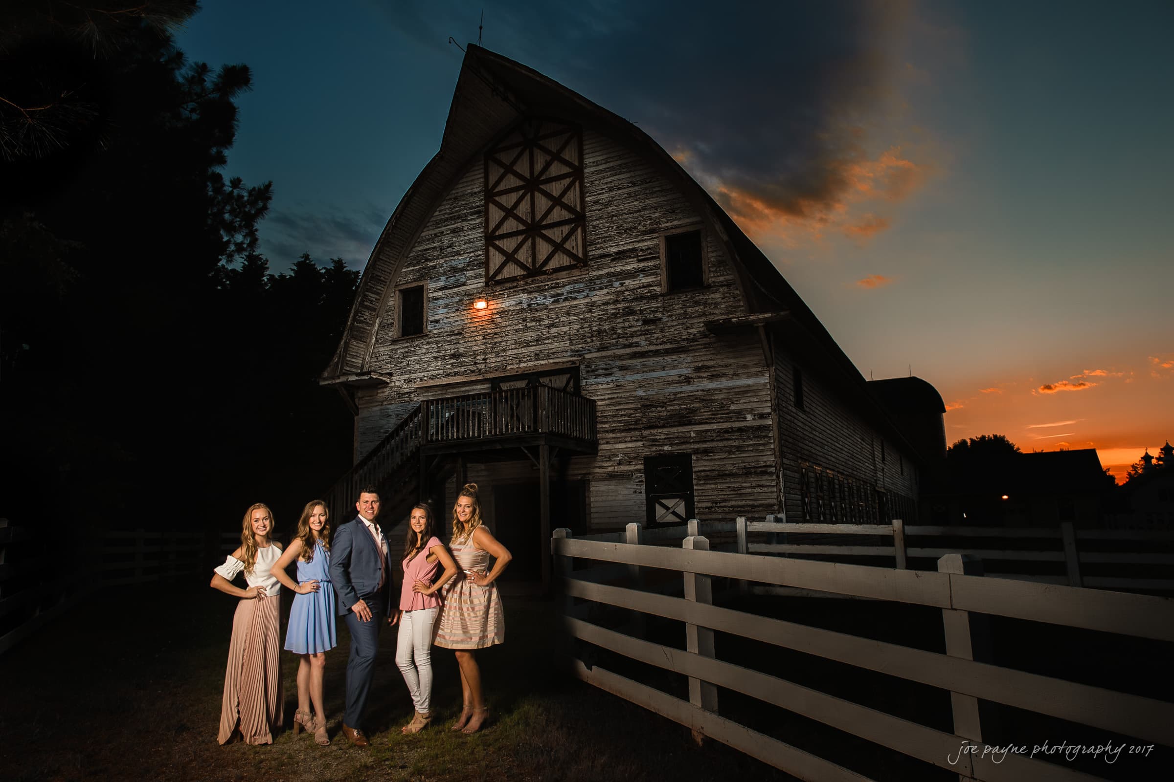 Four people pose in front of a large rustic barn at sunset. The sky is colorful with hues of orange and purple. They are standing on a grassy area bordered by a white fence, casting soft shadows on the ground.