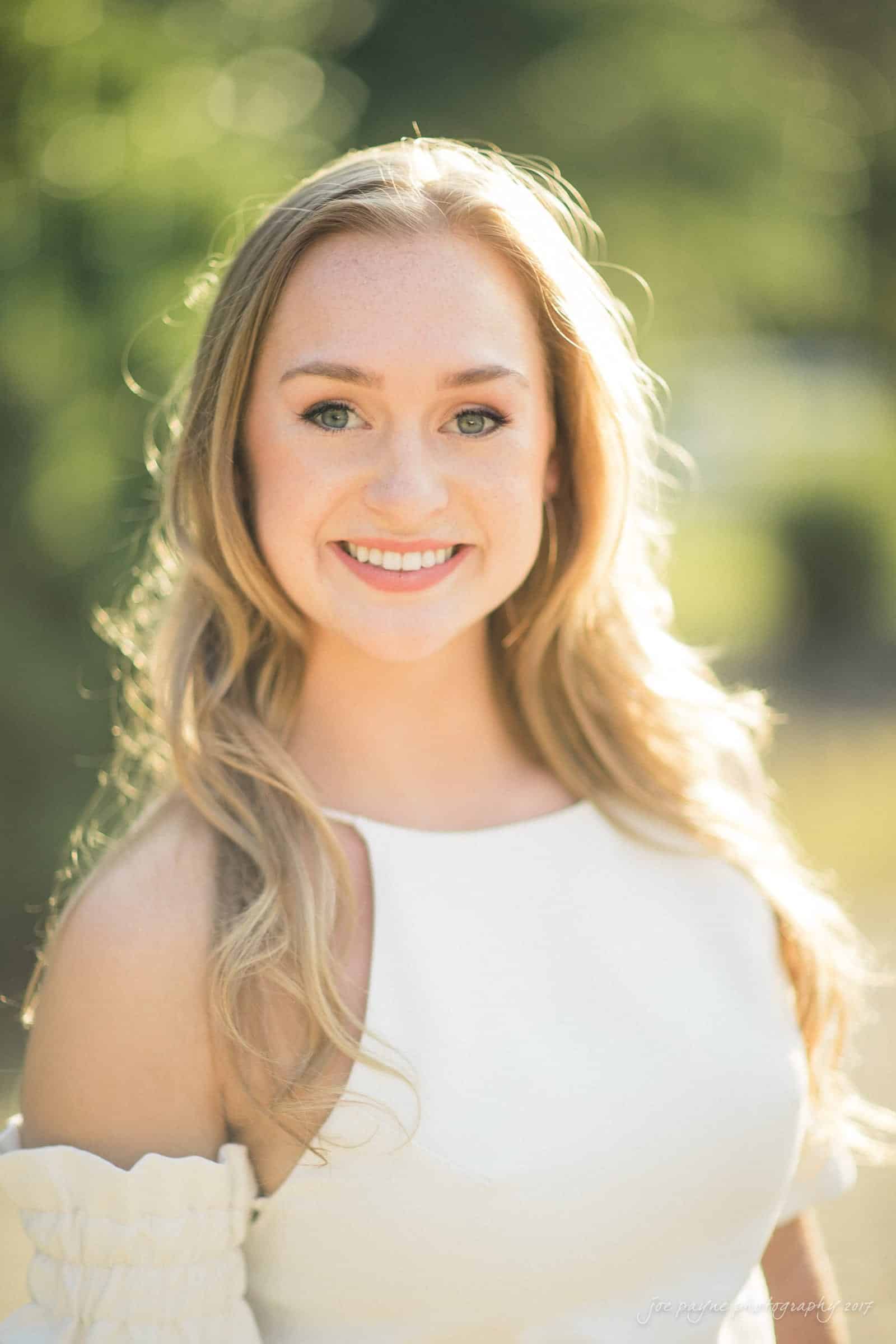 A young woman with long, blonde hair smiles while standing outdoors. She is wearing a white dress with off-the-shoulder sleeves. The background is blurred greenery, giving a soft and natural setting.