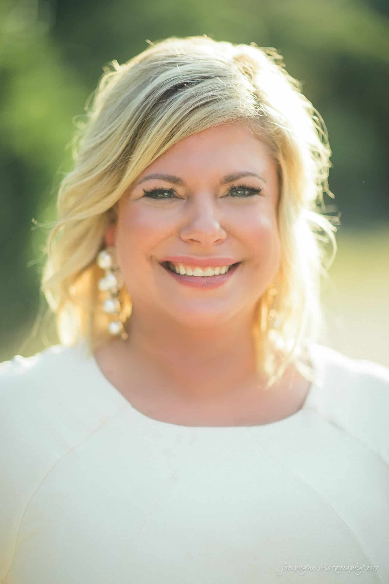 A smiling person with shoulder-length blonde hair, wearing large earrings and a light-colored top, is outdoors in natural sunlight with a blurred green background.