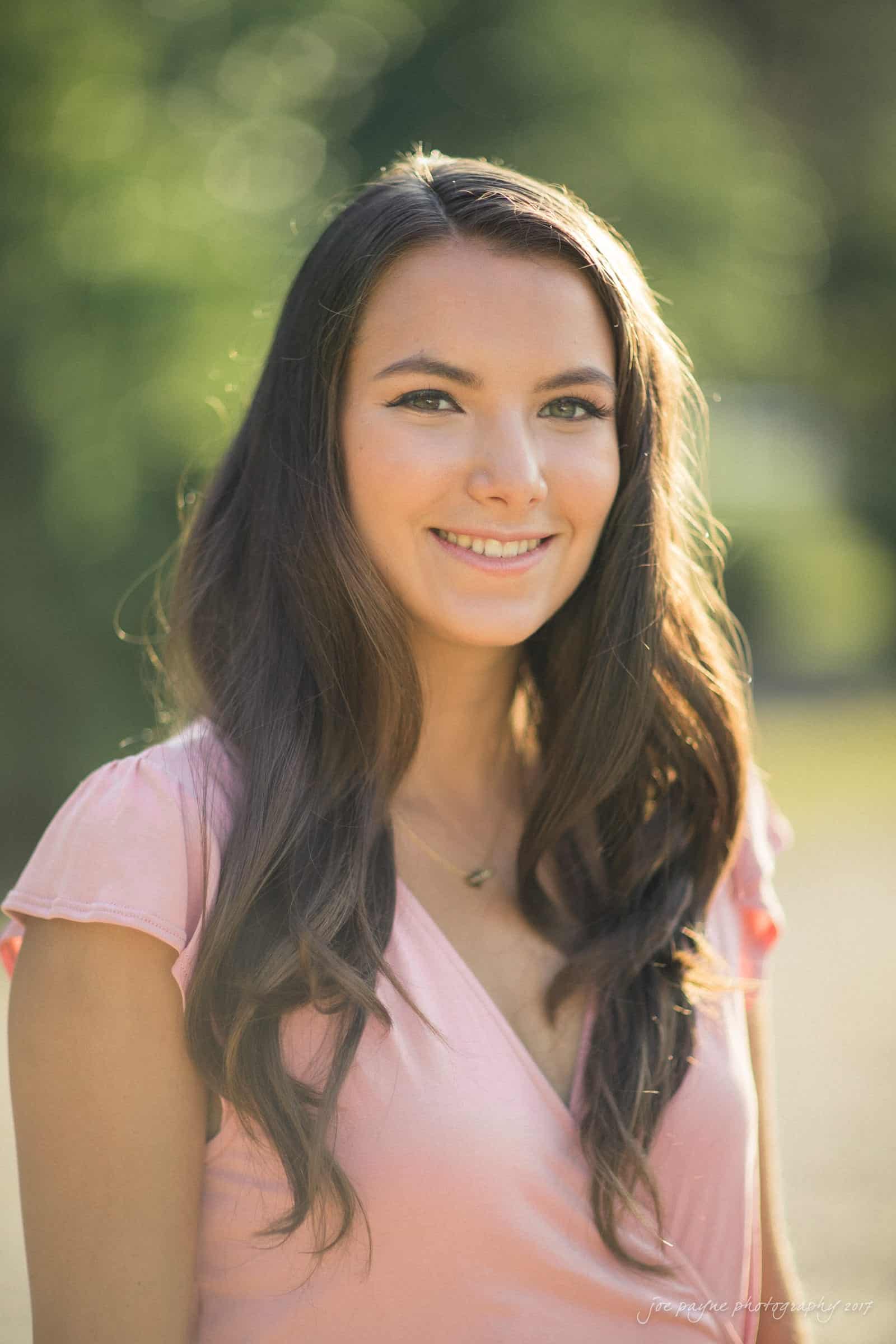A woman with long brown hair and a pink dress smiles softly against a blurred outdoor background. The sunlight softly illuminates her face and hair.