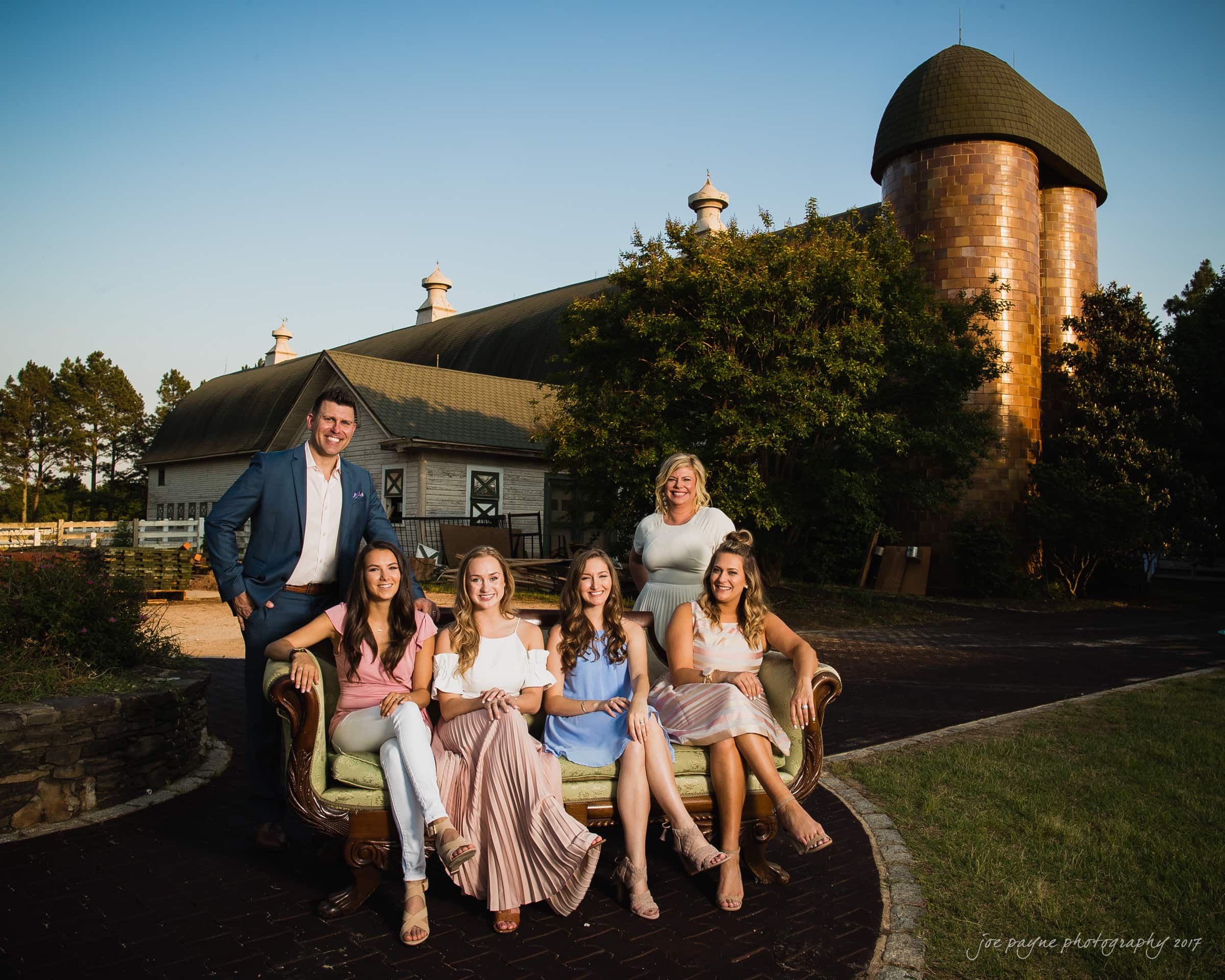 A group photo with six people sitting on an antique sofa outdoors. Behind them is a large barn with a silo. The setting suggests a rural landscape in sunny weather. Five women and one man are smiling for the camera.