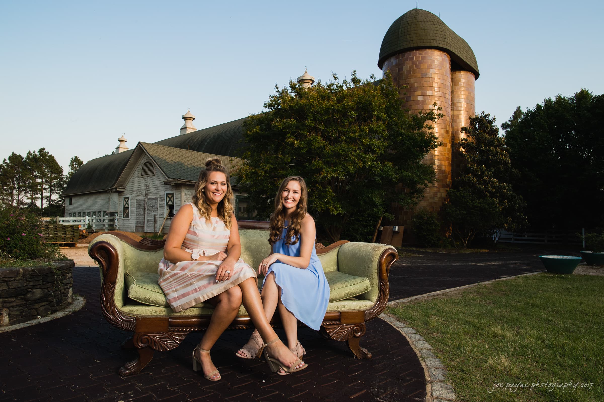 Two women sitting on a vintage couch outdoors, with a rustic barn and silo in the background. Both are smiling and dressed in summer dresses, one striped and the other blue. The setting is peaceful with trees and a clear sky.