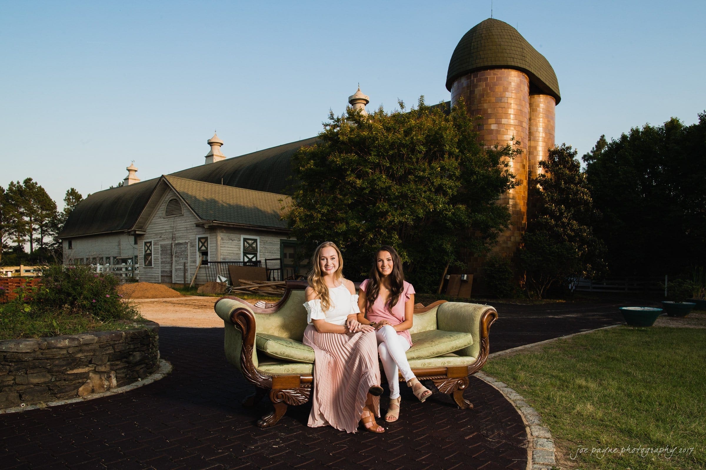 Two women are sitting on a vintage green couch outdoors. They are smiling and holding hands. Behind them is a large barn with a silo under a clear blue sky. The surrounding area is paved and grassy.